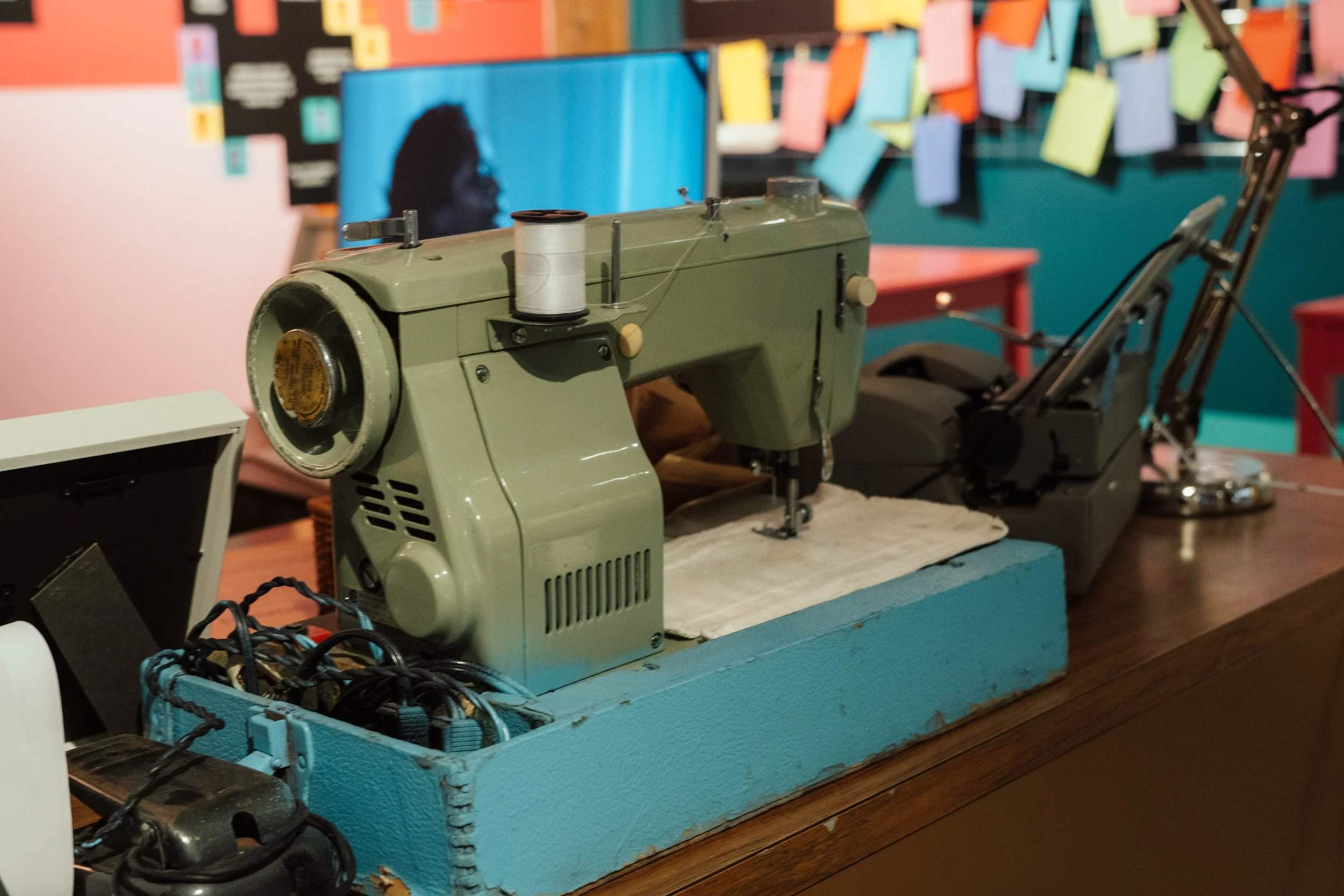 Vintage green sewing machine on a wooden table with a roll of white thread on top, in front of a colorful wall with sticky notes and a TV screen in the background.