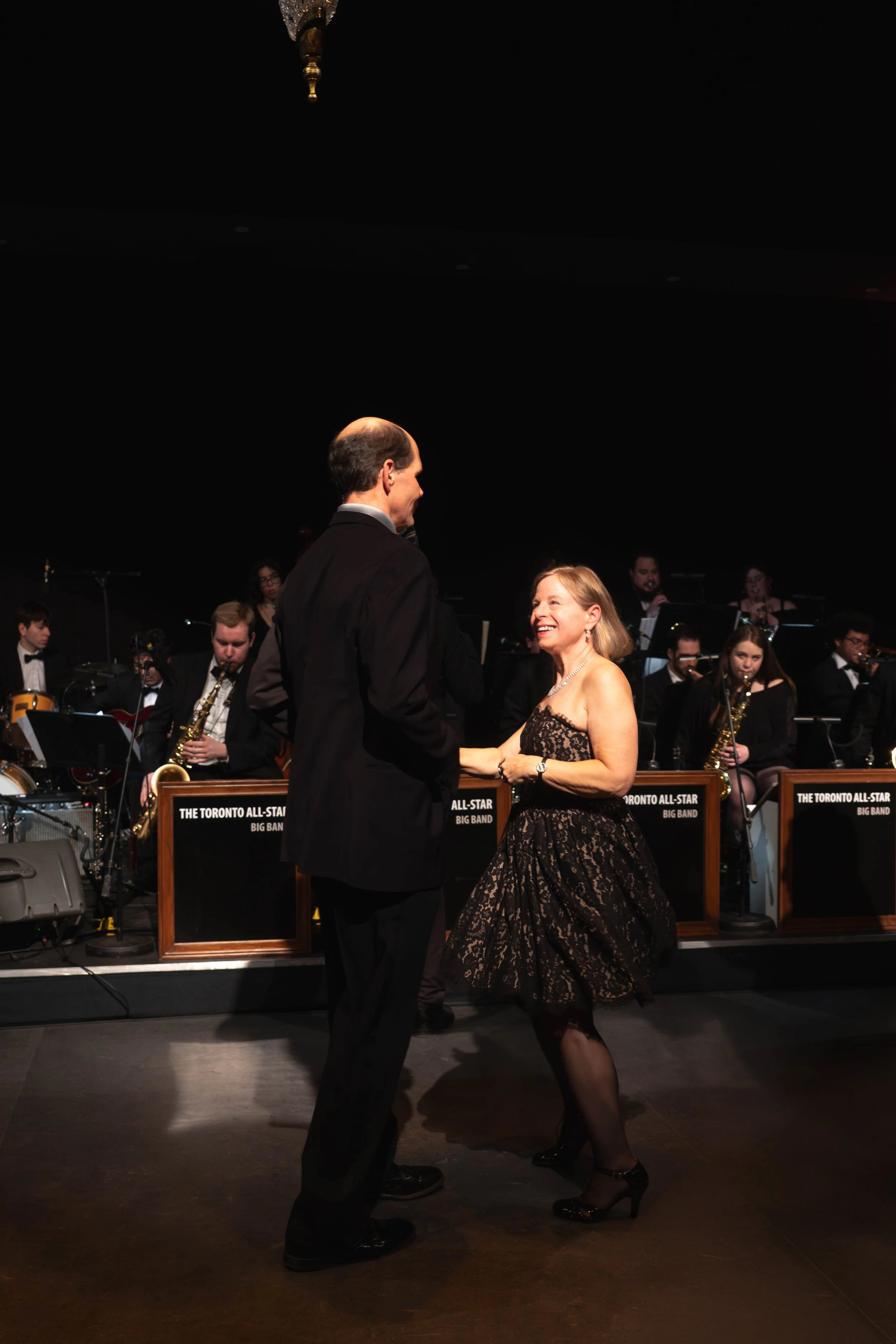 A man and woman dancing together on a dance floor during a live big band music performance. The background has an orchestra with musicians playing instruments, seated behind music stands labeled "The Toronto All-Star Big Band."