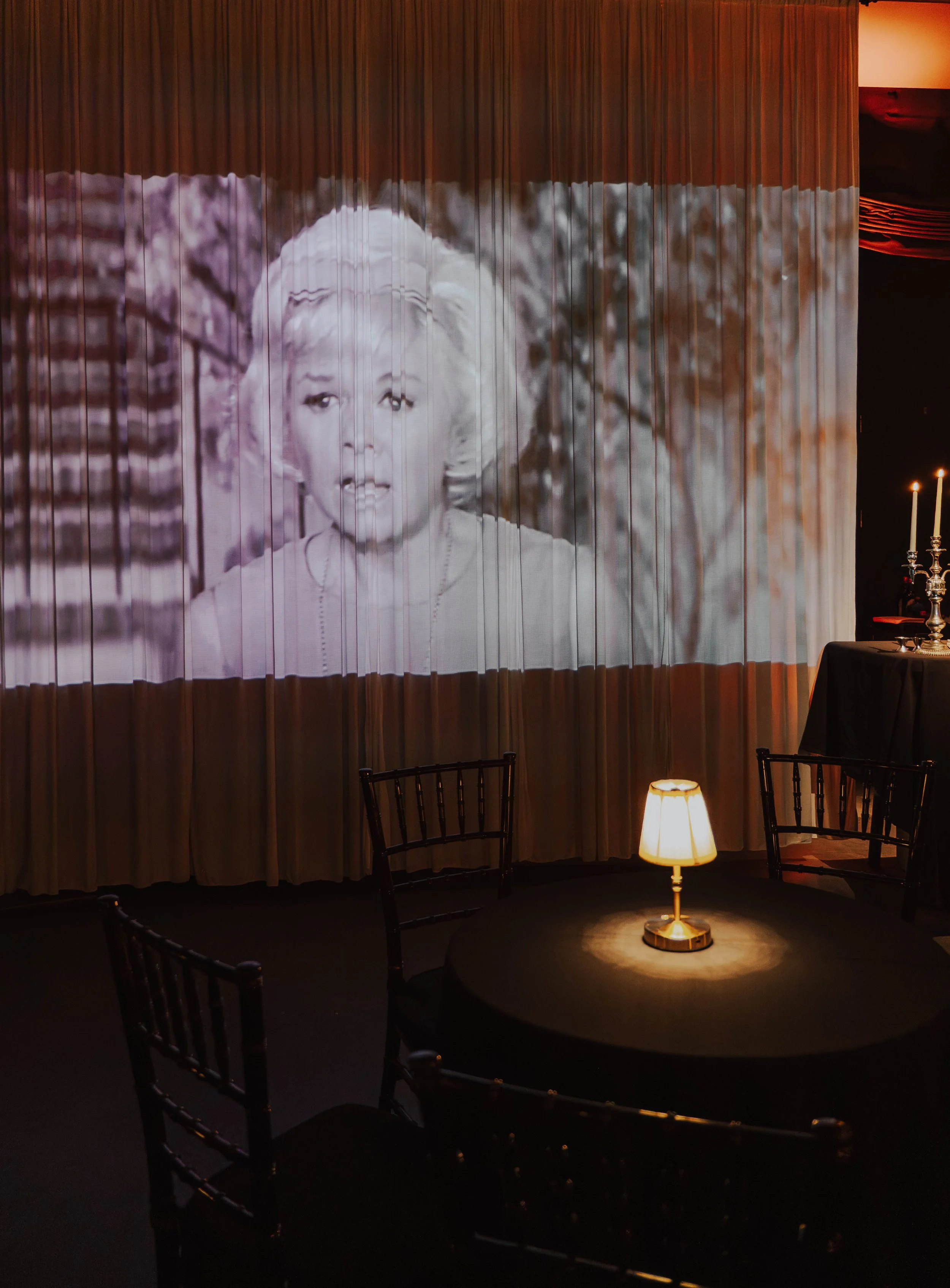 A black and white photograph of Marilyn Monroe projected on a curtain with chairs, a small lamp, and a table with candles in the foreground.