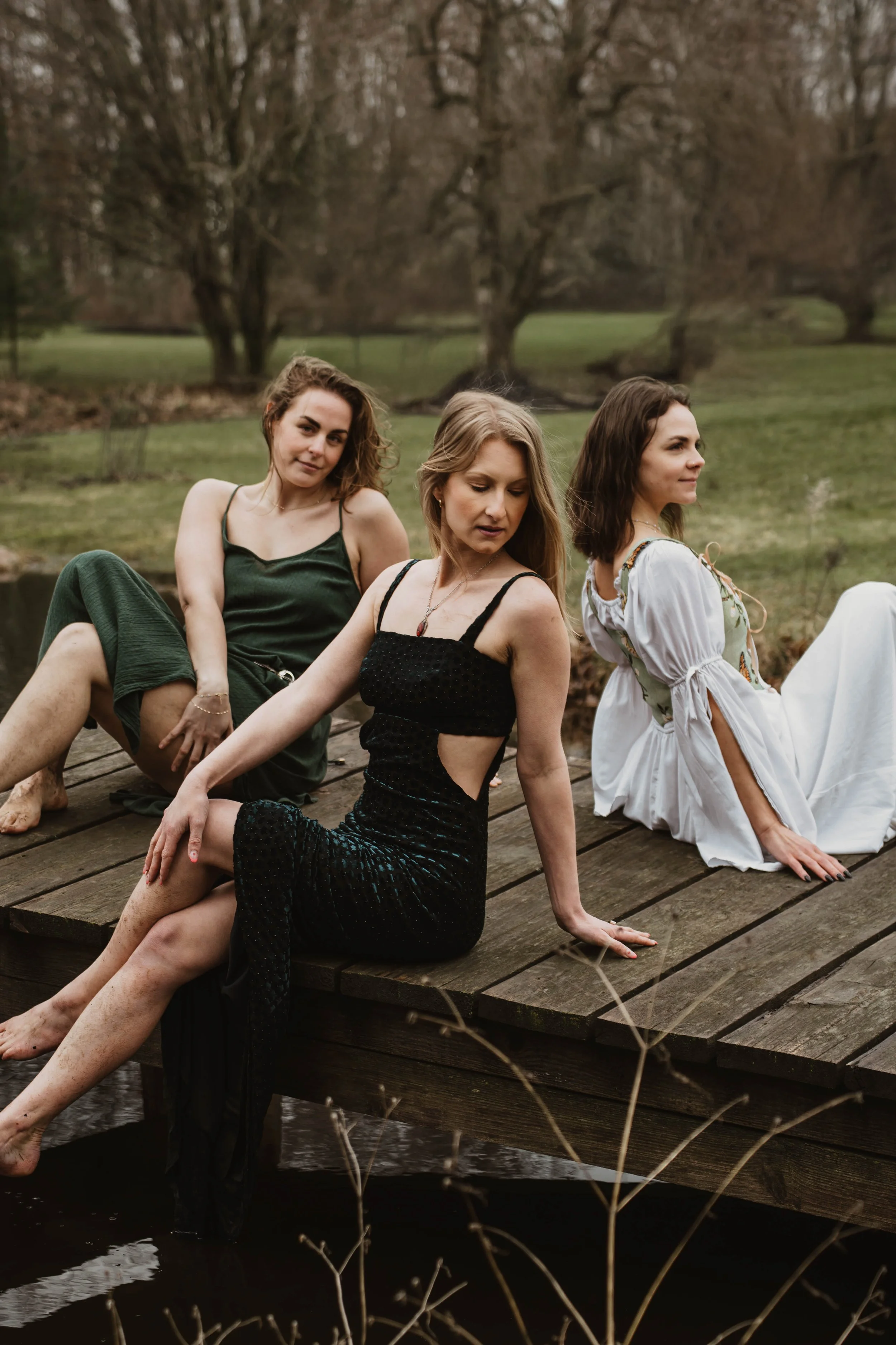Three women sitting on a wooden dock near a pond outdoors in a natural setting during daytime.