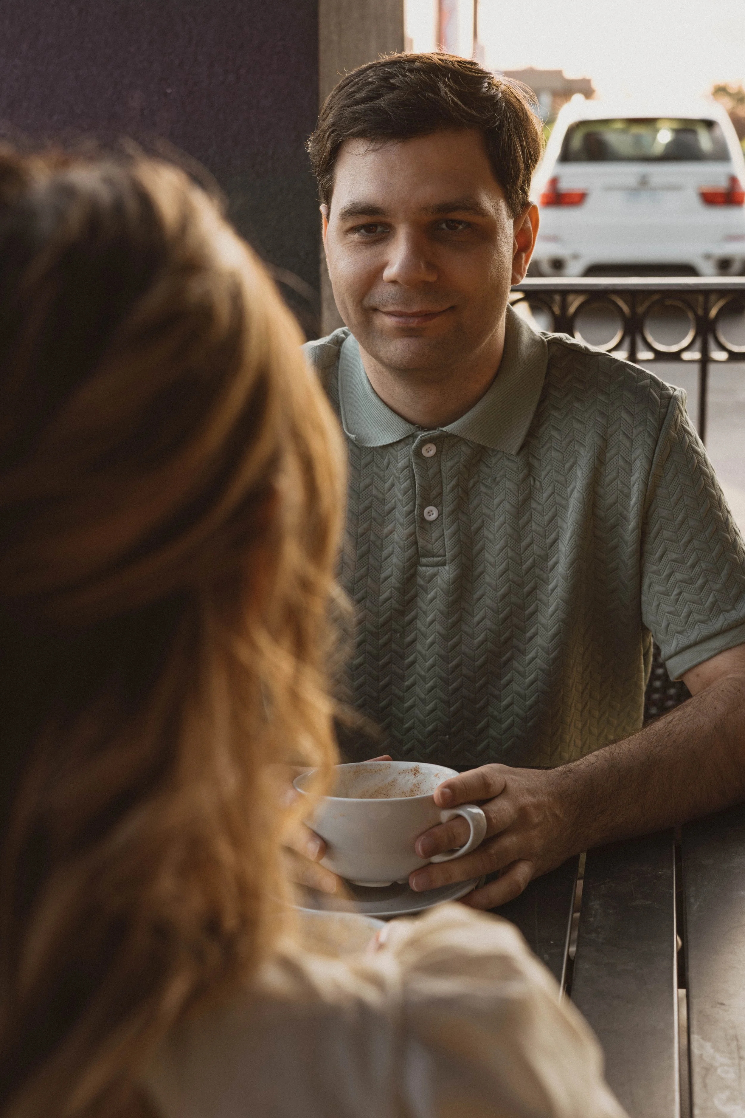 A man with dark hair, wearing a patterned polo shirt, holding a white bowl, possibly with food, and smiling slightly, engaged in conversation with a woman with light brown or blonde hair, seen from behind, at an outdoor cafe or restaurant during suns