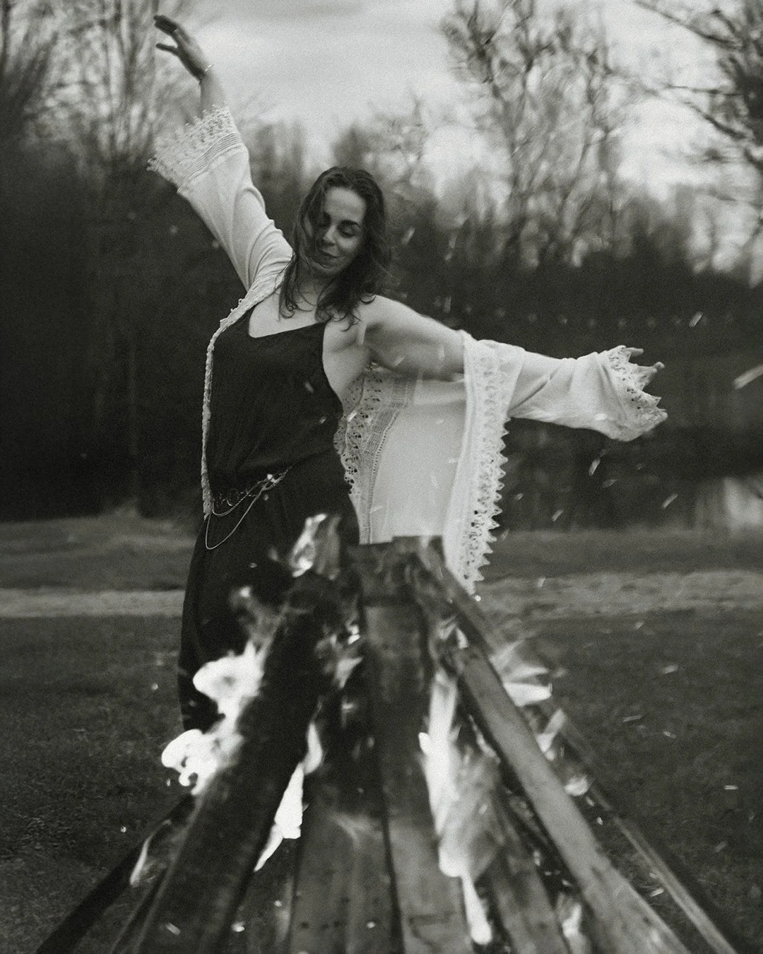 A woman dancing outdoors by a pond, wearing a black dress and a light shawl with lace edges, as leaves fall around her.