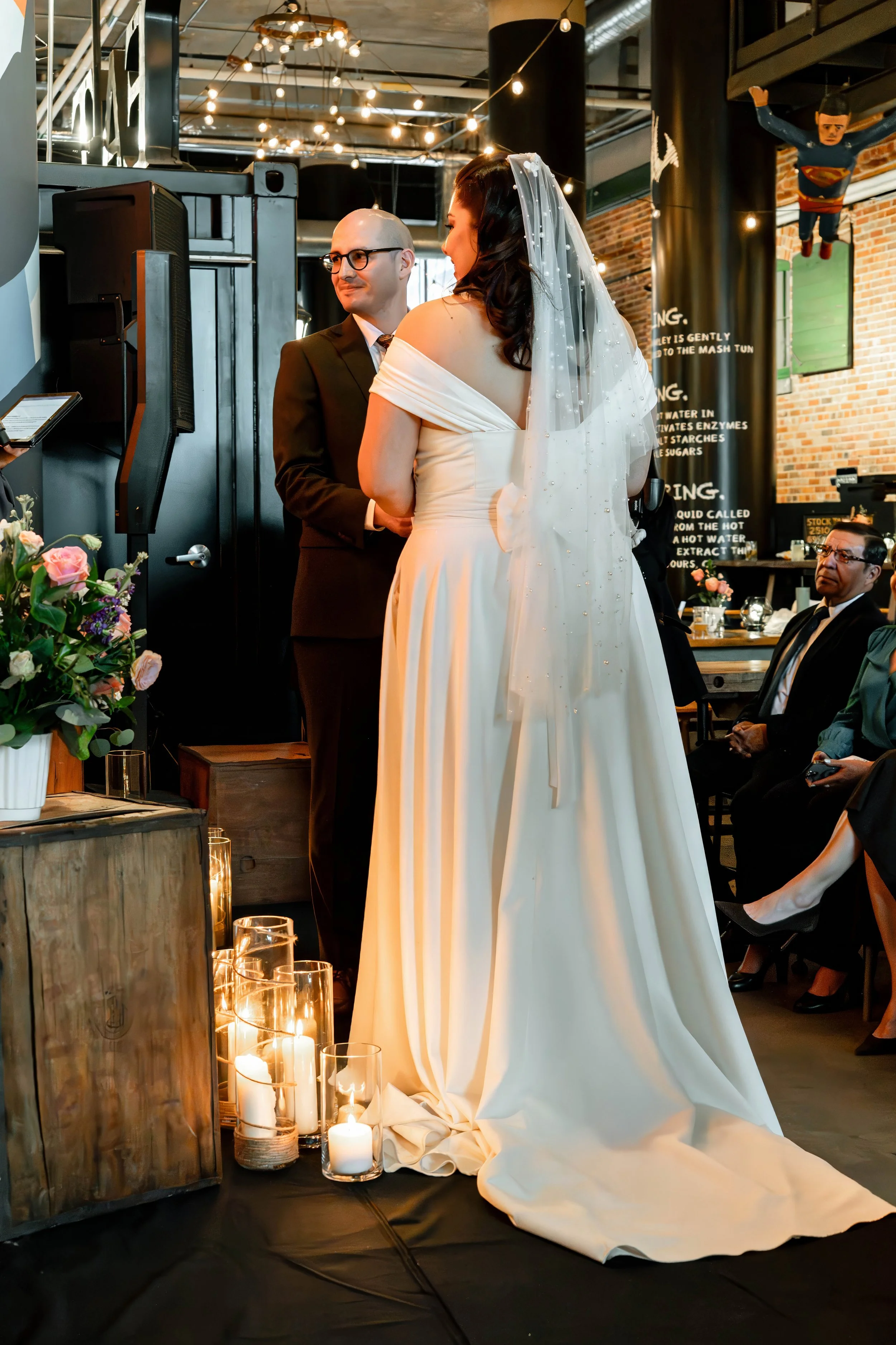 A bride and groom standing at their wedding ceremony in a cozy, industrial-style venue with exposed brick walls, string lights, and black pillars. The bride is wearing a white wedding gown with a veil, and the groom is dressed in a black suit with gl