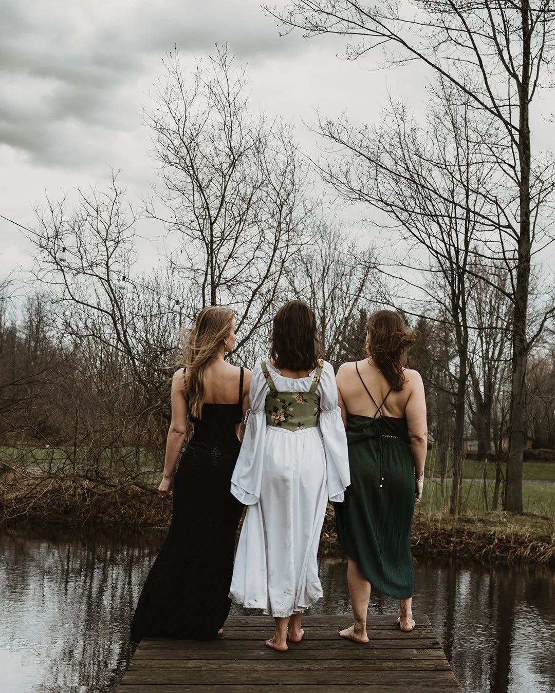 Three women with long hair, standing barefoot on a wooden dock by a river, facing away, under leafless trees on an overcast day.