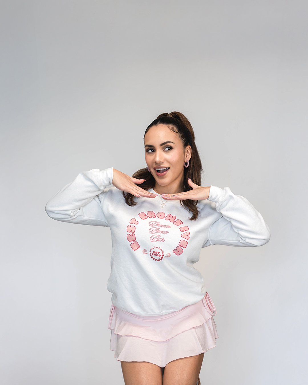 A young woman with dark hair in a high ponytail, wearing a white sweatshirt and pink layered shorts, posing against a plain gray background with a playful expression and hand gestures near her face.