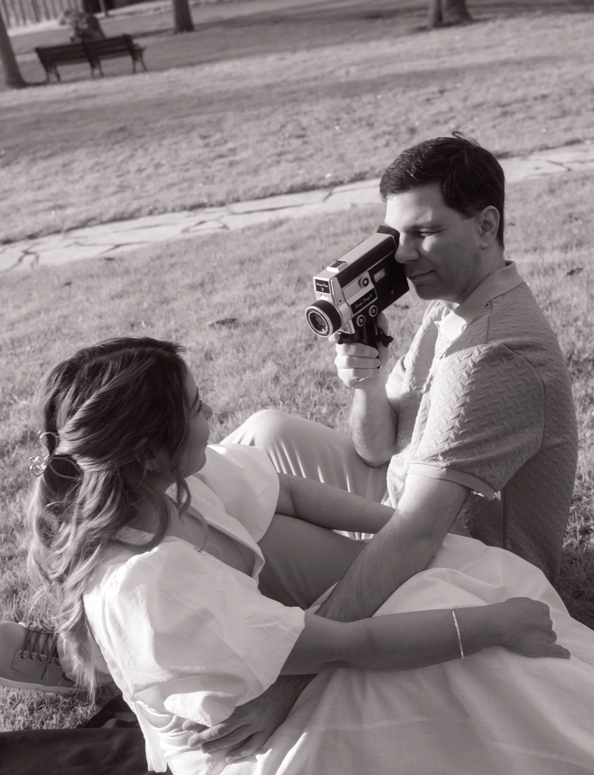 A man holding a vintage movie camera outdoors, with a woman sitting in front of him, in a park with trees and benches in the background.