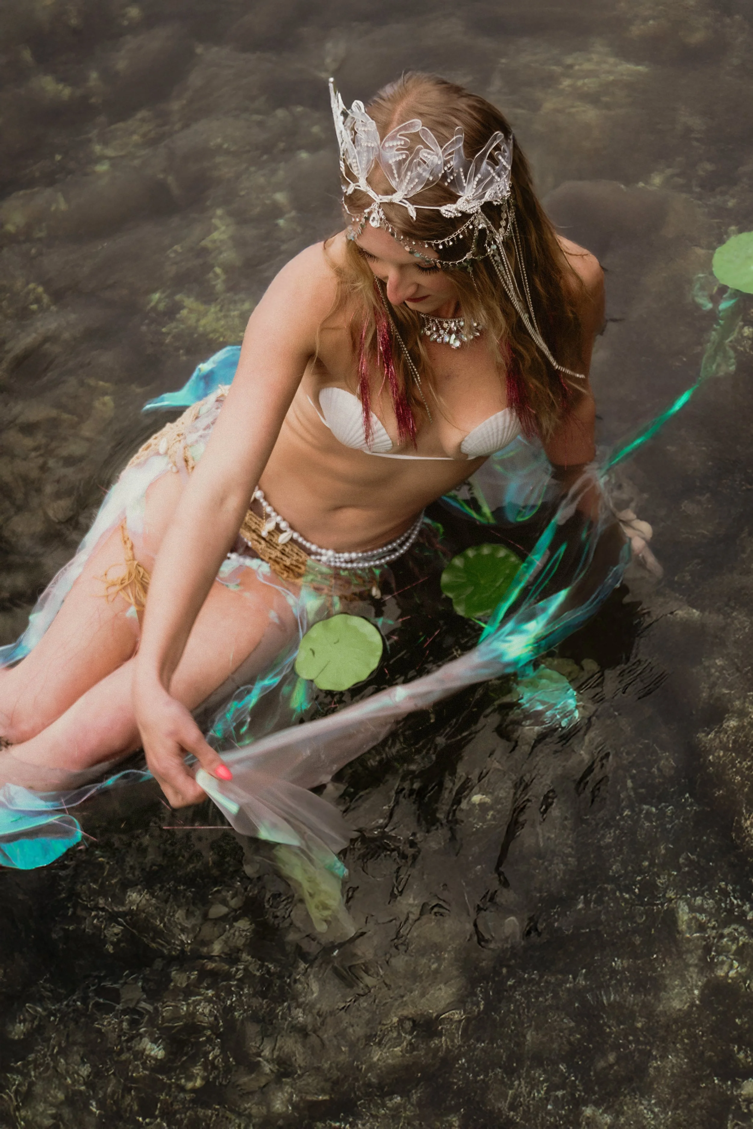 A woman wearing a decorative butterfly headpiece and jewelry sitting in a shallow body of water with lily pads, holding a transparent umbrella in a natural outdoor setting.