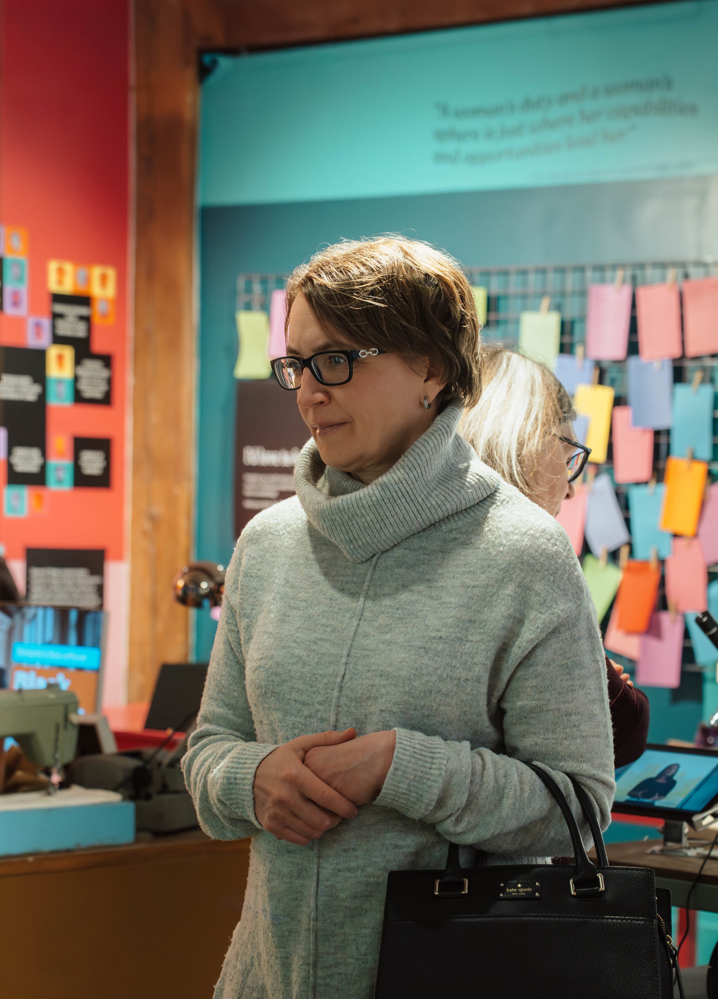 A woman wearing glasses and a light gray turtleneck sweater, holding a black handbag, stands indoors near a colorful display of notes and aboard with info in the background.