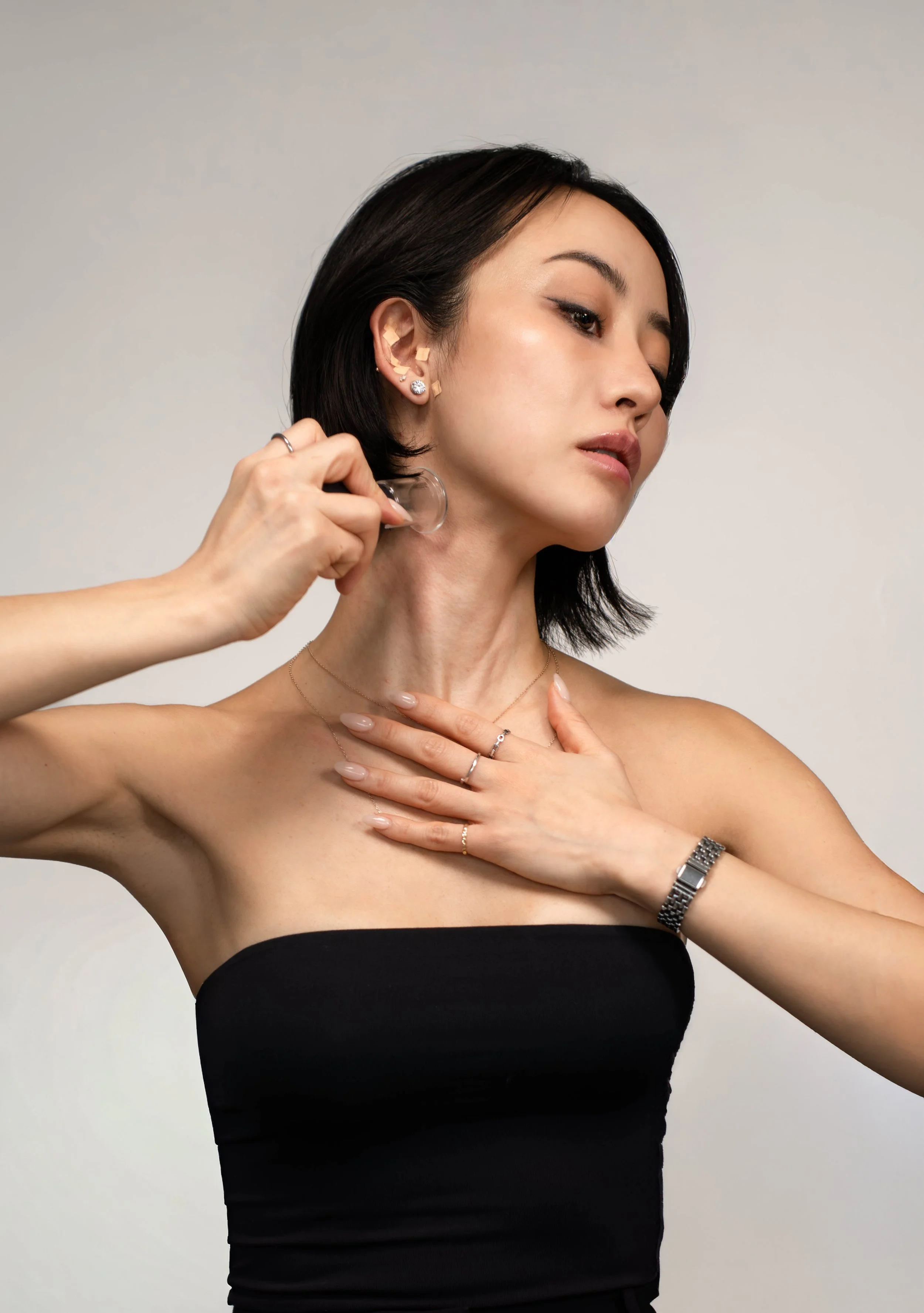 A woman with short black hair applying perfume behind her ear while touching her chest, wearing a strapless black top and jewelry.