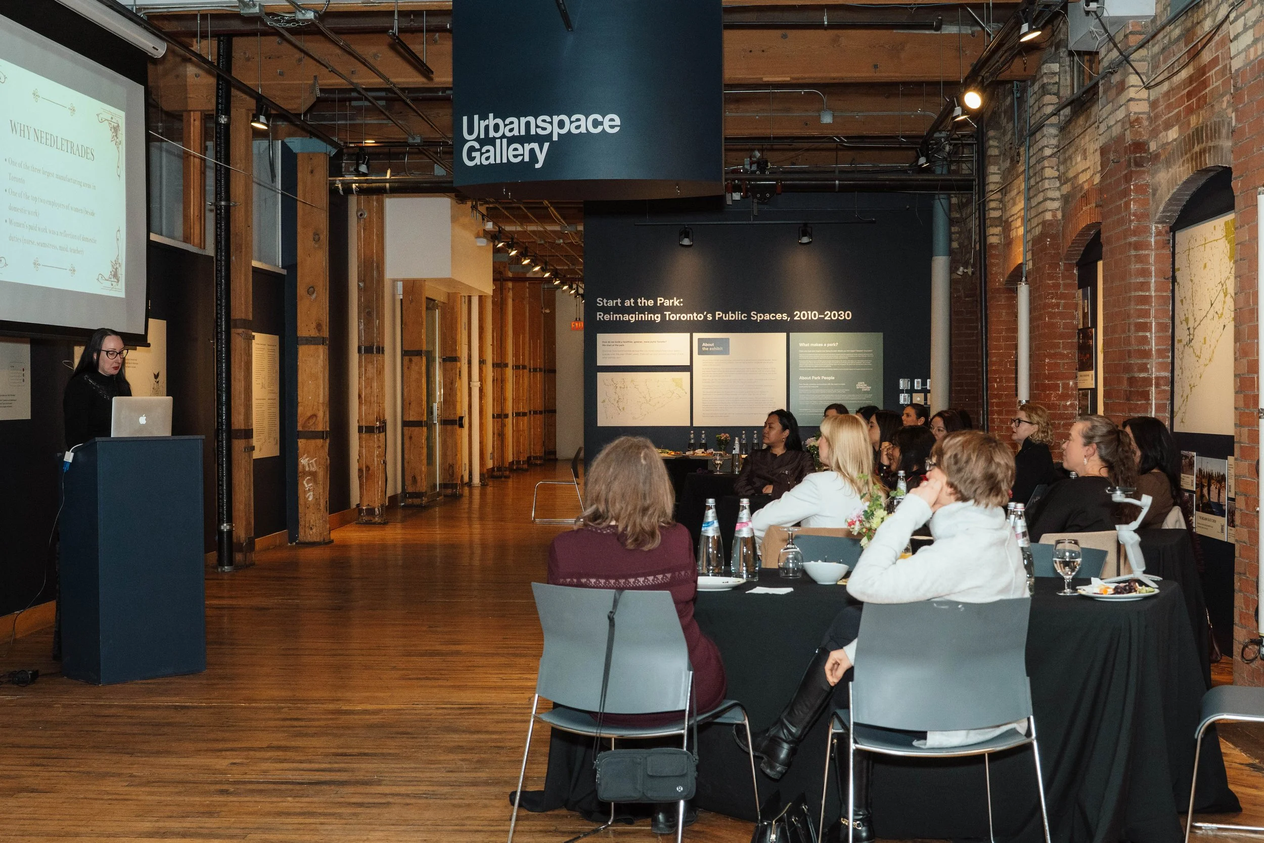 A woman giving a presentation to an audience in a gallery space with wooden floors and brick walls, titled 'Urbanspace Gallery'. Audience members are seated at tables with water bottles and glasses.