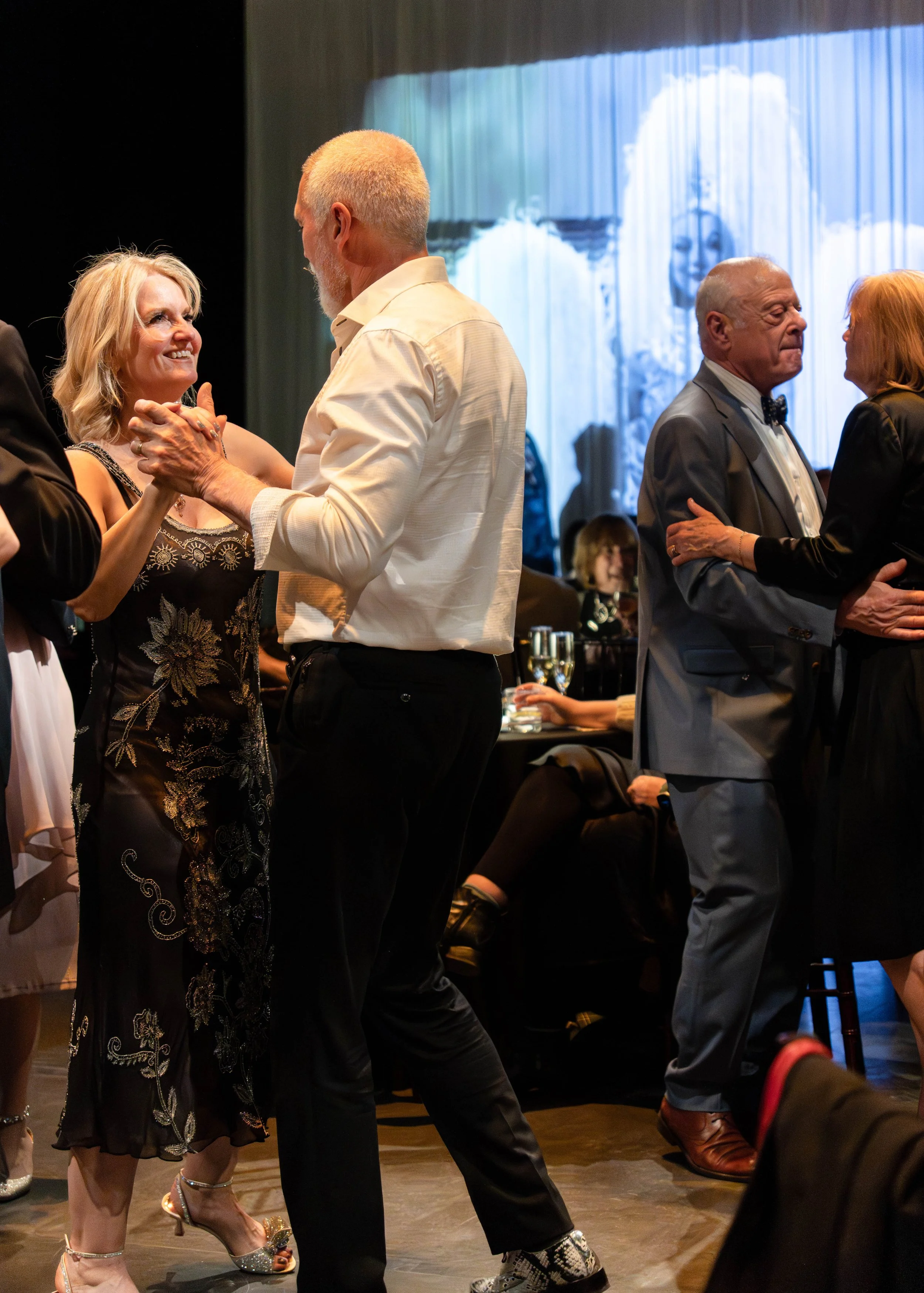 Couples dancing at a formal event, with a woman in a black embroidered dress and a man in a white shirt dancing in the foreground, a couple in tuxedos dancing in the background, and a projection of a woman with a white wig on the screen behind them.