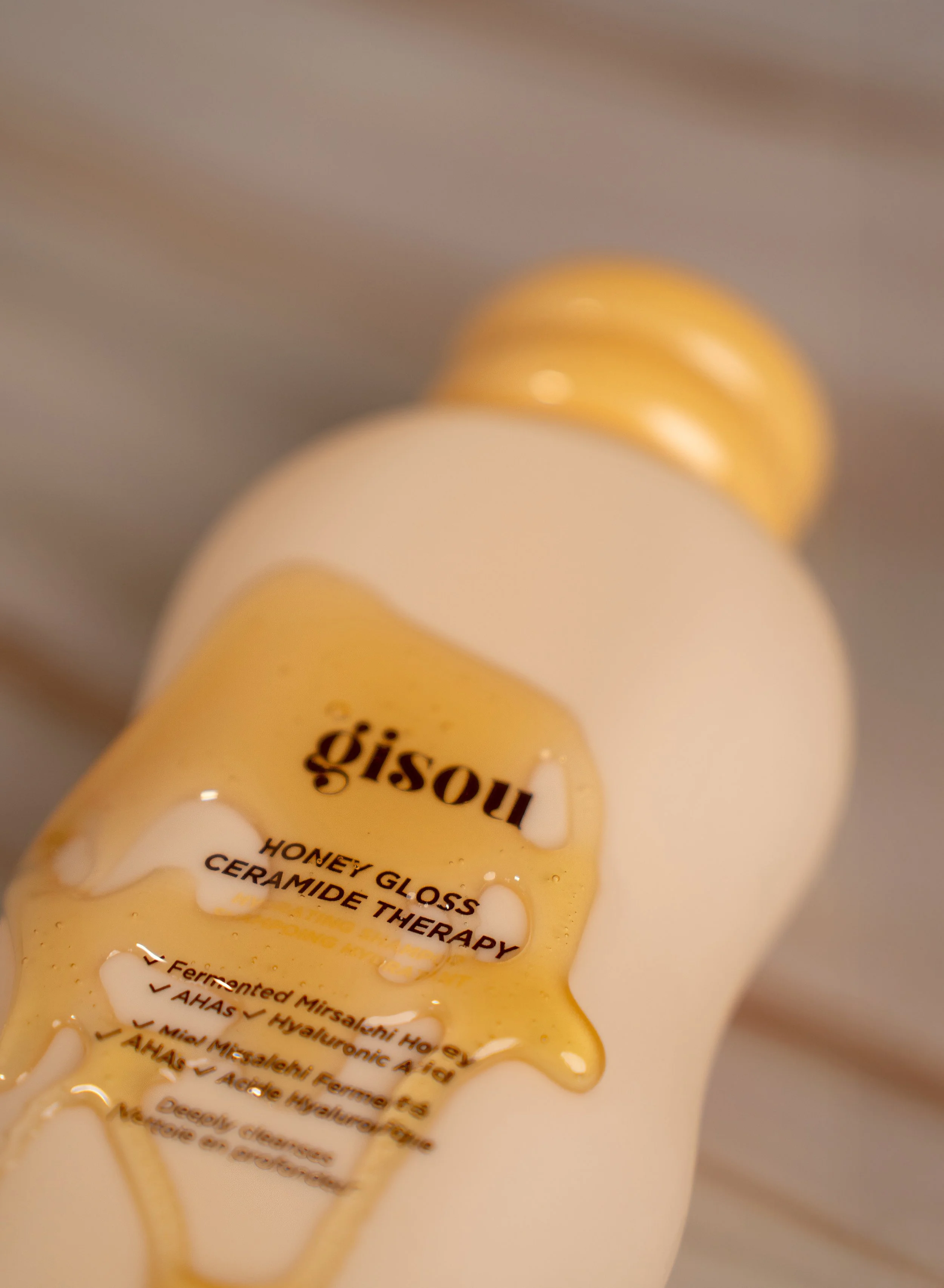 Close-up of a white bottle with a yellow-honey-colored label, titled 'Gisou Honey Gloss' with 'Ceramide Therapy' written underneath, and honey dripping from the label.