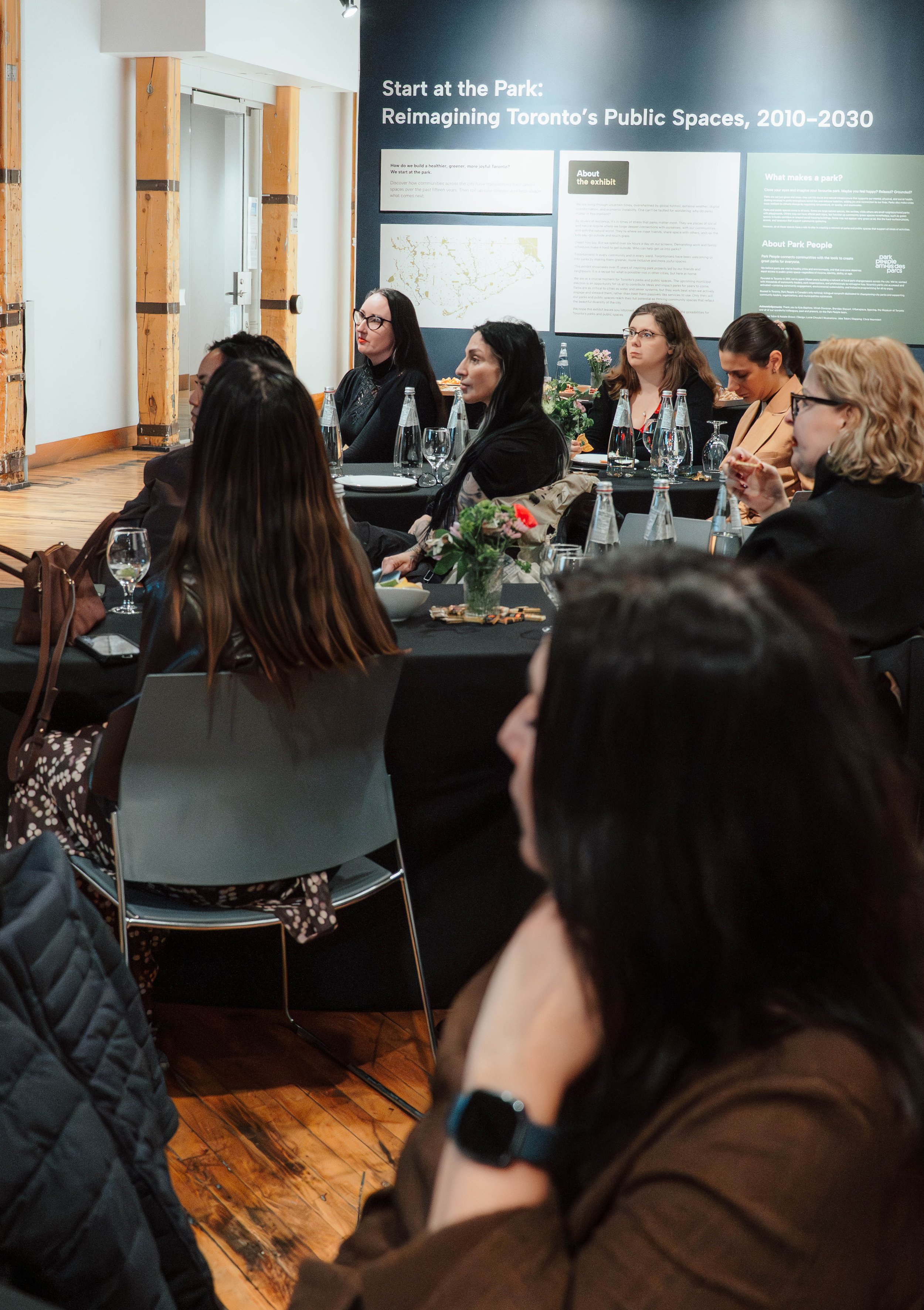 A group of women sitting at a conference table in a museum or gallery, listening to a presentation on reimagining Toronto's public spaces from 2010 to 2030.