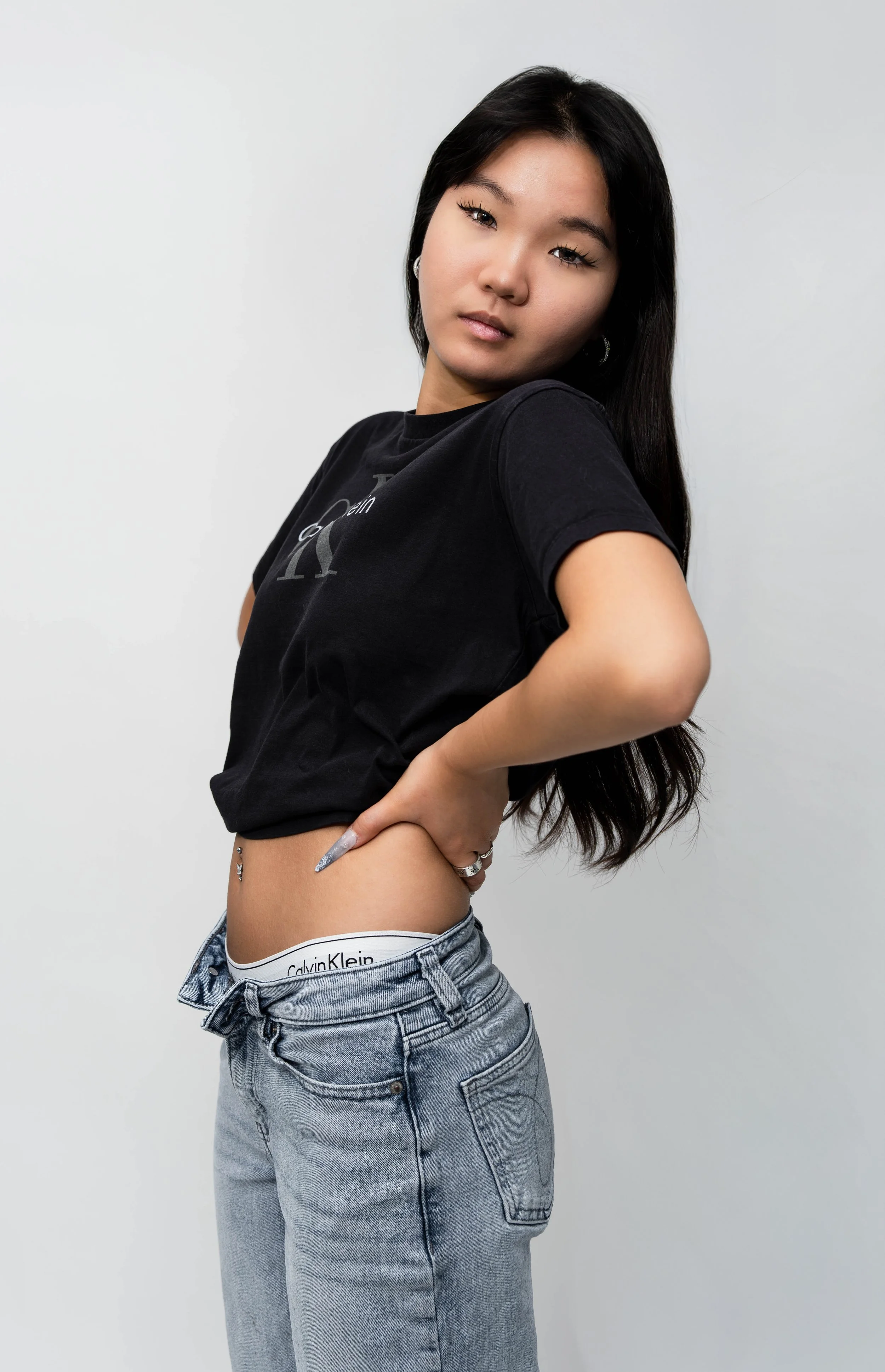 Young woman with long dark hair posing against a plain white background, wearing a black Calvin Klein t-shirt, light-wash jeans, and visible Calvin Klein underwear waistband.