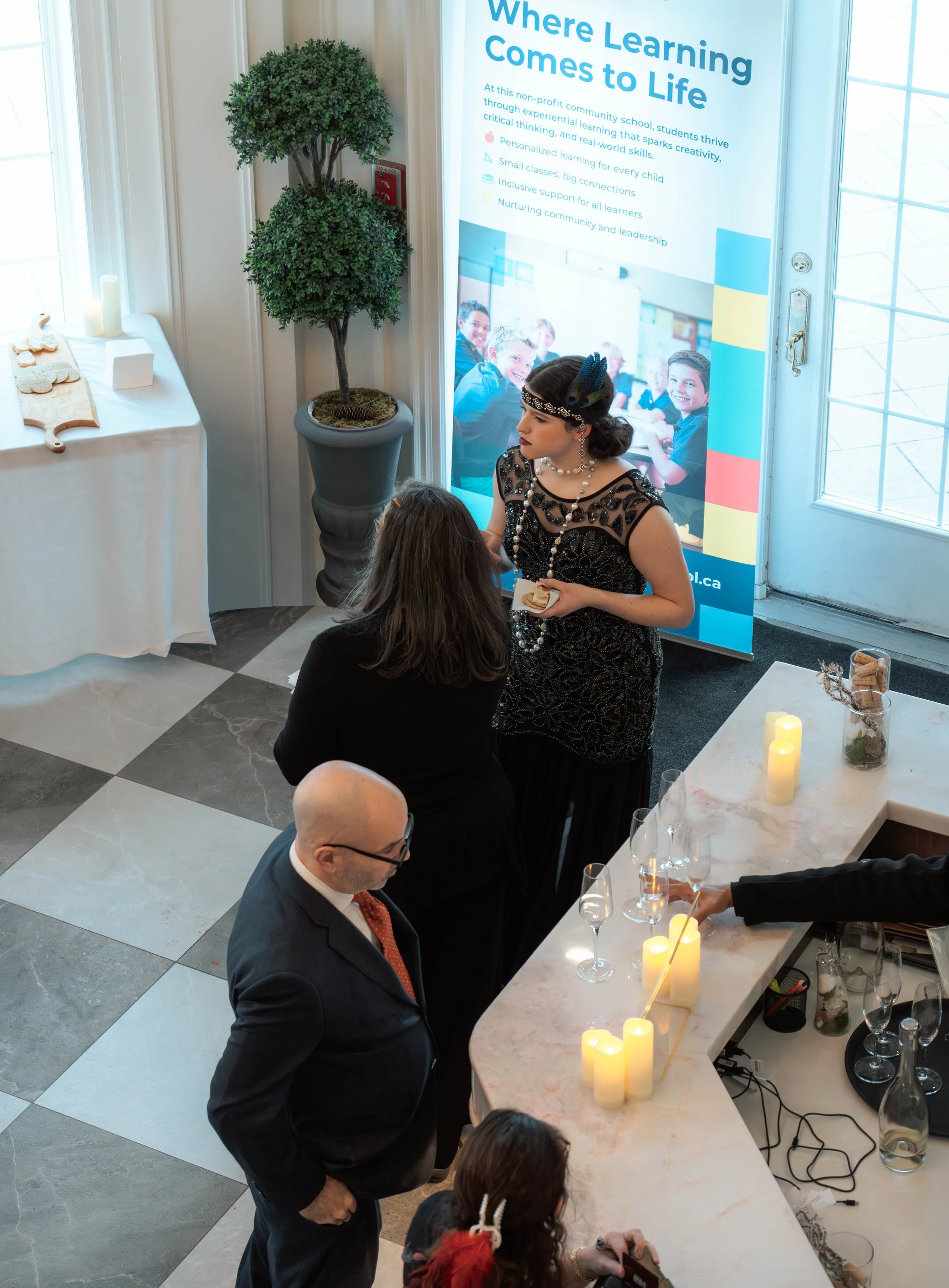 A woman dressed in black vintage attire with pearls and a headband is talking to another woman at a social event near a bar counter with lit candles and glasses. Two men are nearby, one with glasses and a suit, and a young girl with hair accessories.