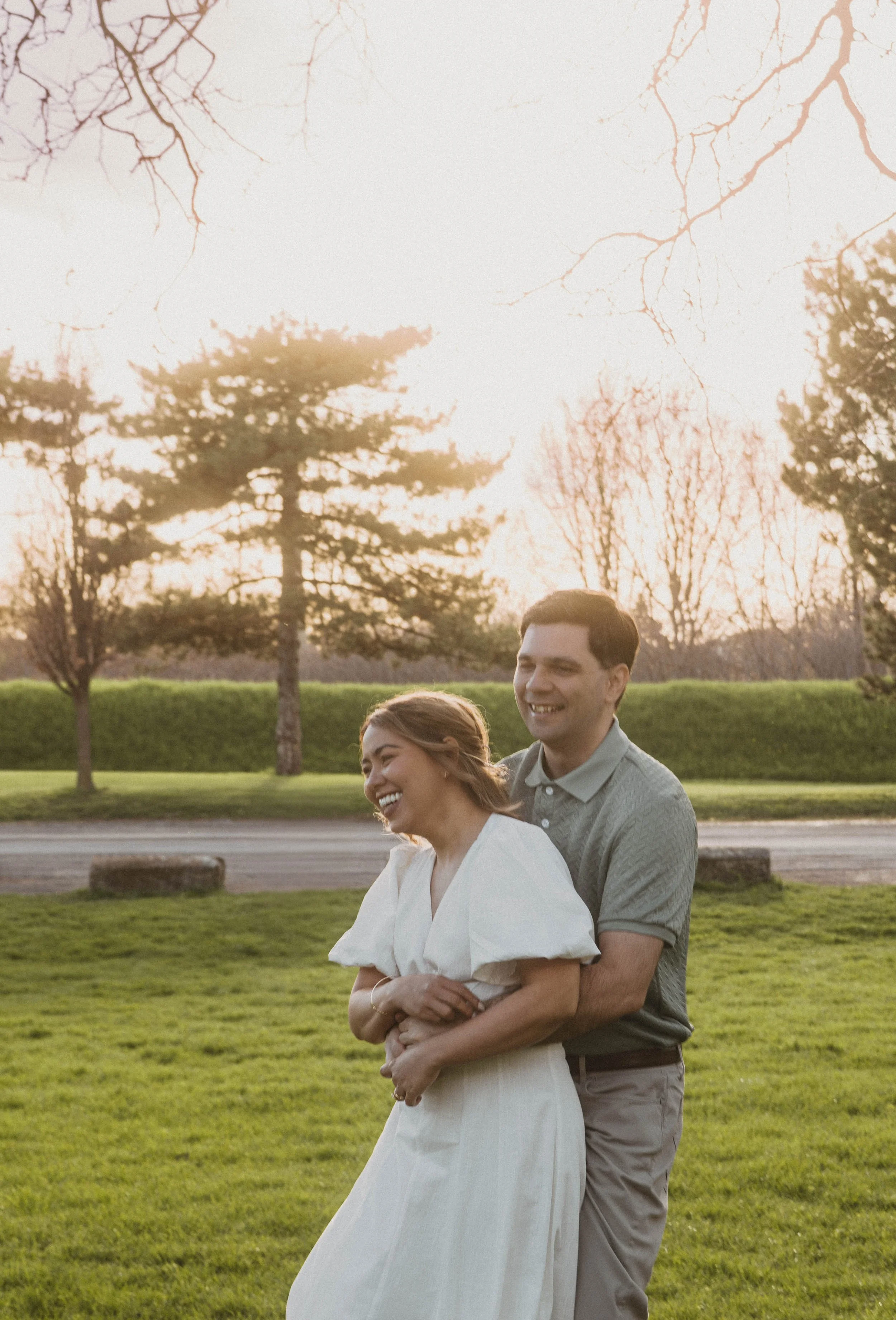 A happy couple, a woman in a white dress and a man in a gray shirt, are embracing outdoors in a park during sunset, with trees and a hedge in the background.