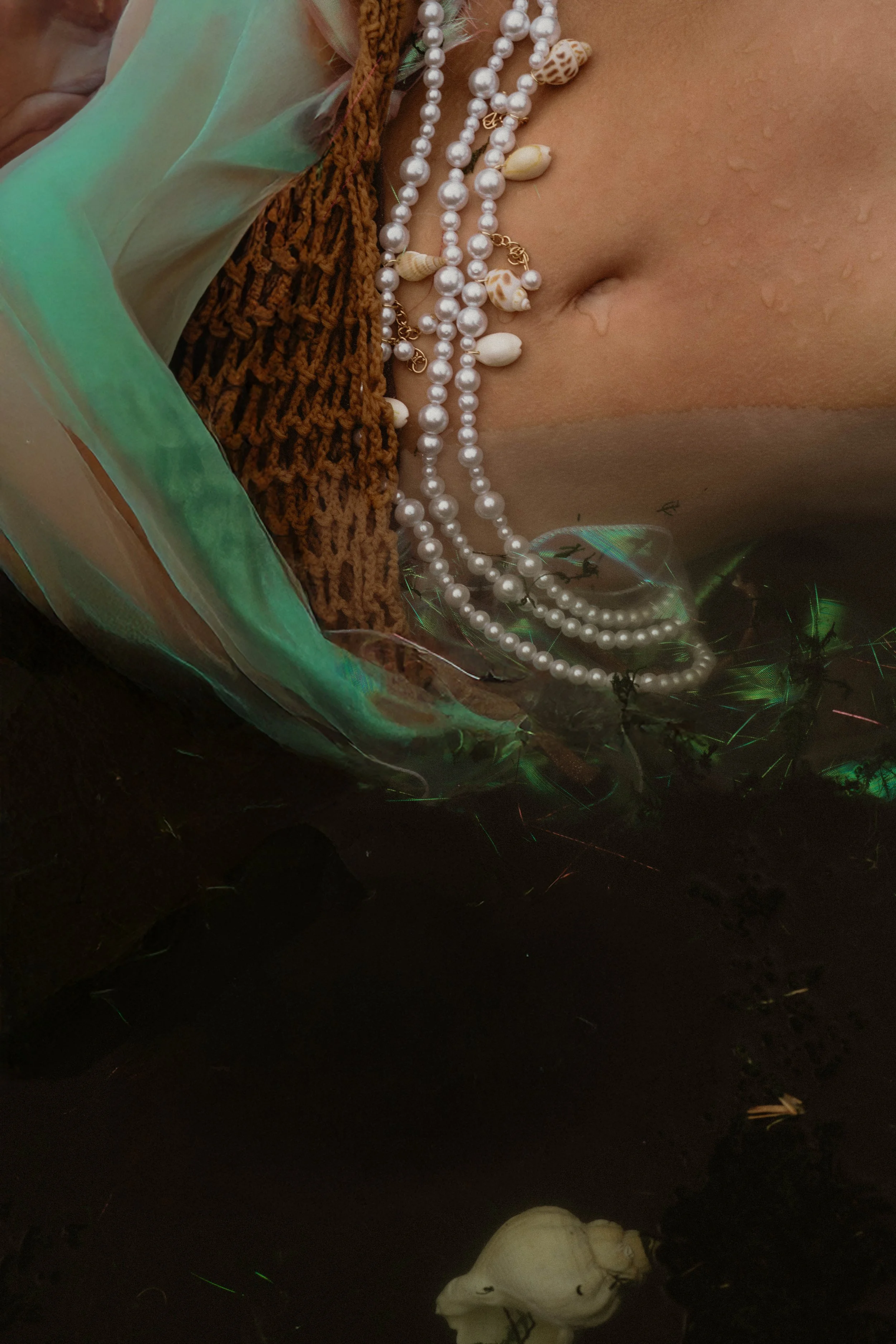 Close-up of a person's neck and upper chest adorned with multiple pearl necklaces and seashell pendants, with a brown knit garment and translucent greenish fabric around the shoulders, and water droplets on the skin.