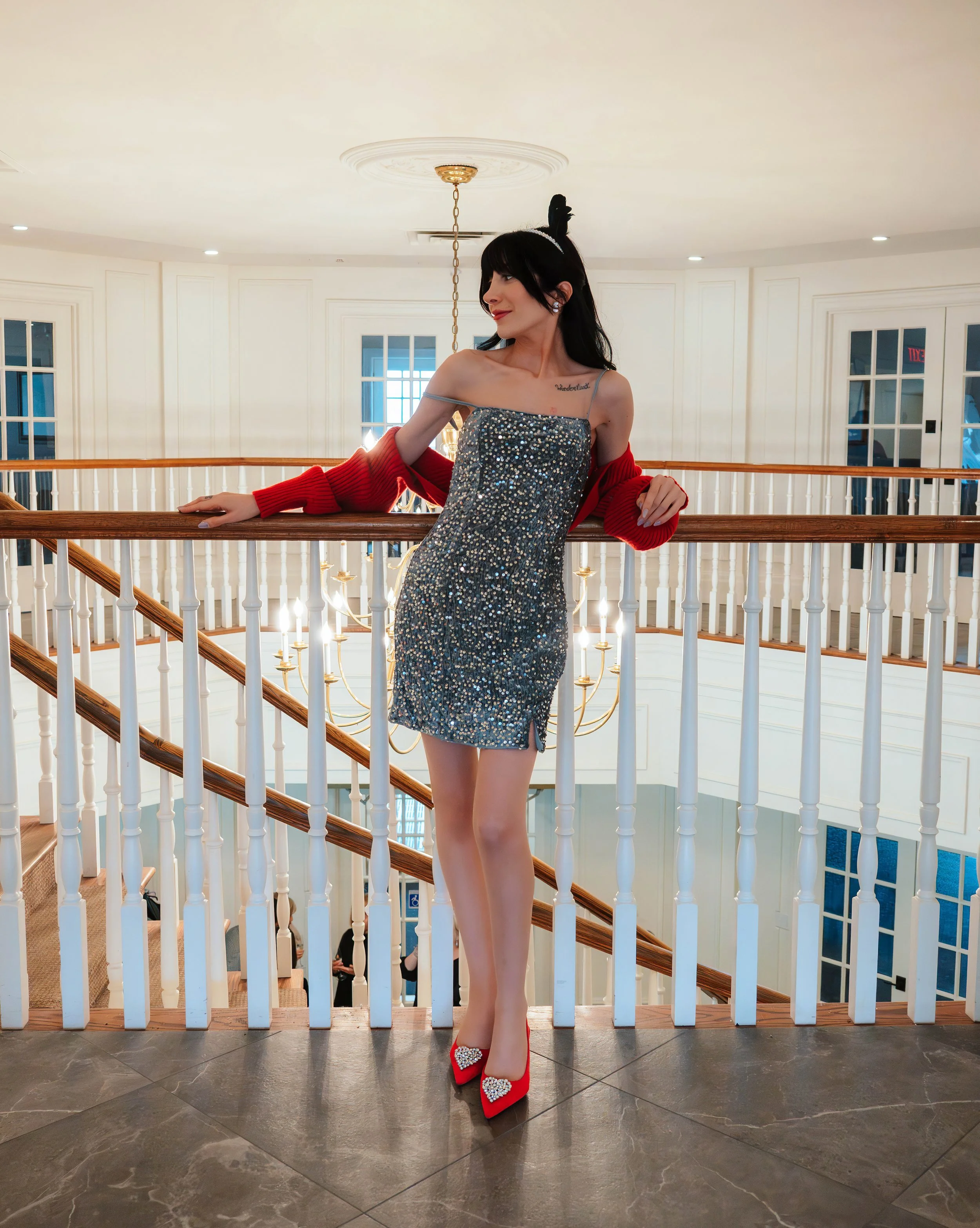 A woman in a glittery silver dress and red high heels with a heart design stands on a staircase landing in a well-lit, white interior space, with her arms resting on a wooden railing.