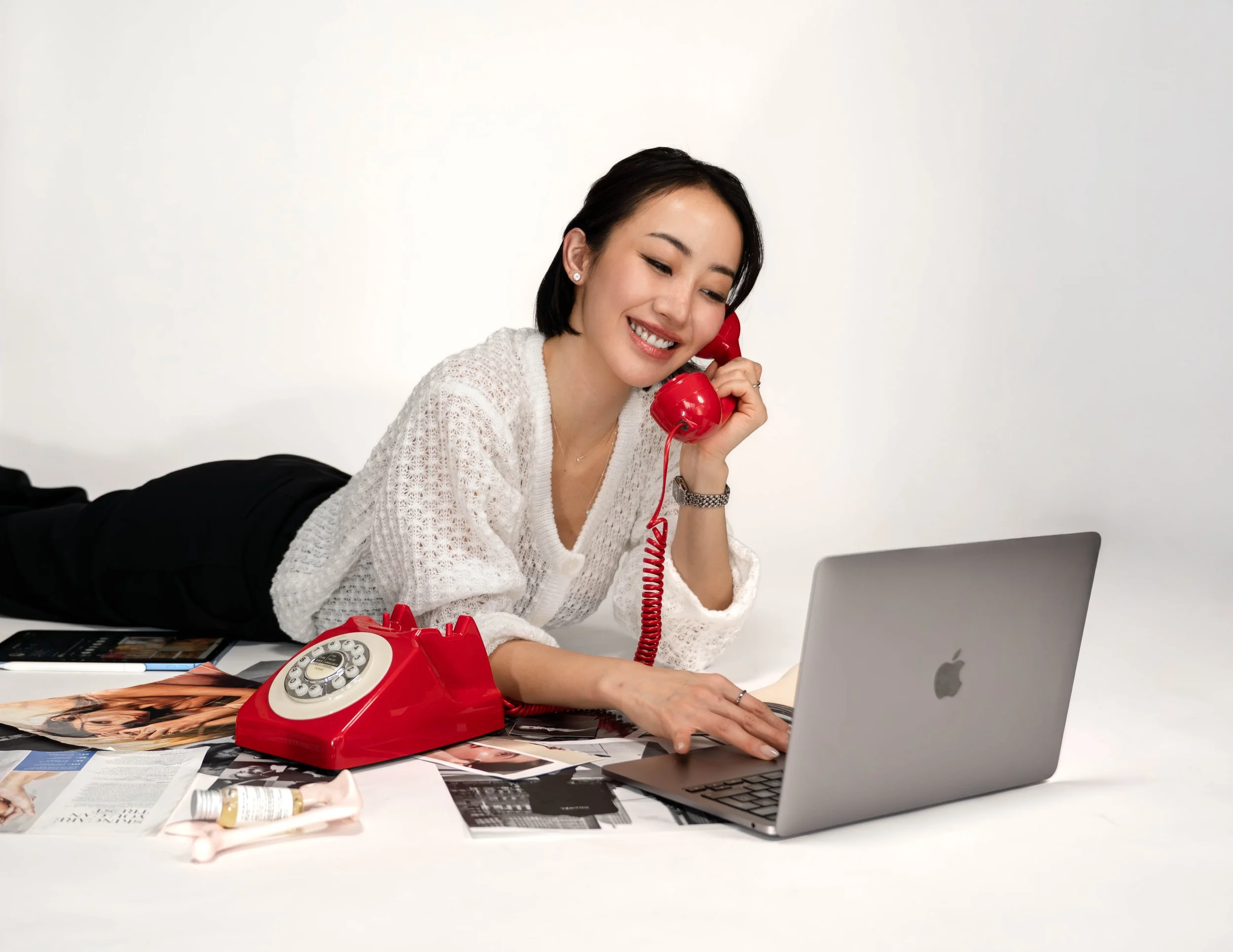 Woman lying on her stomach on a white surface, talking on a red vintage rotary phone, using a silver MacBook, surrounded by magazines, a bottle, and a calculator