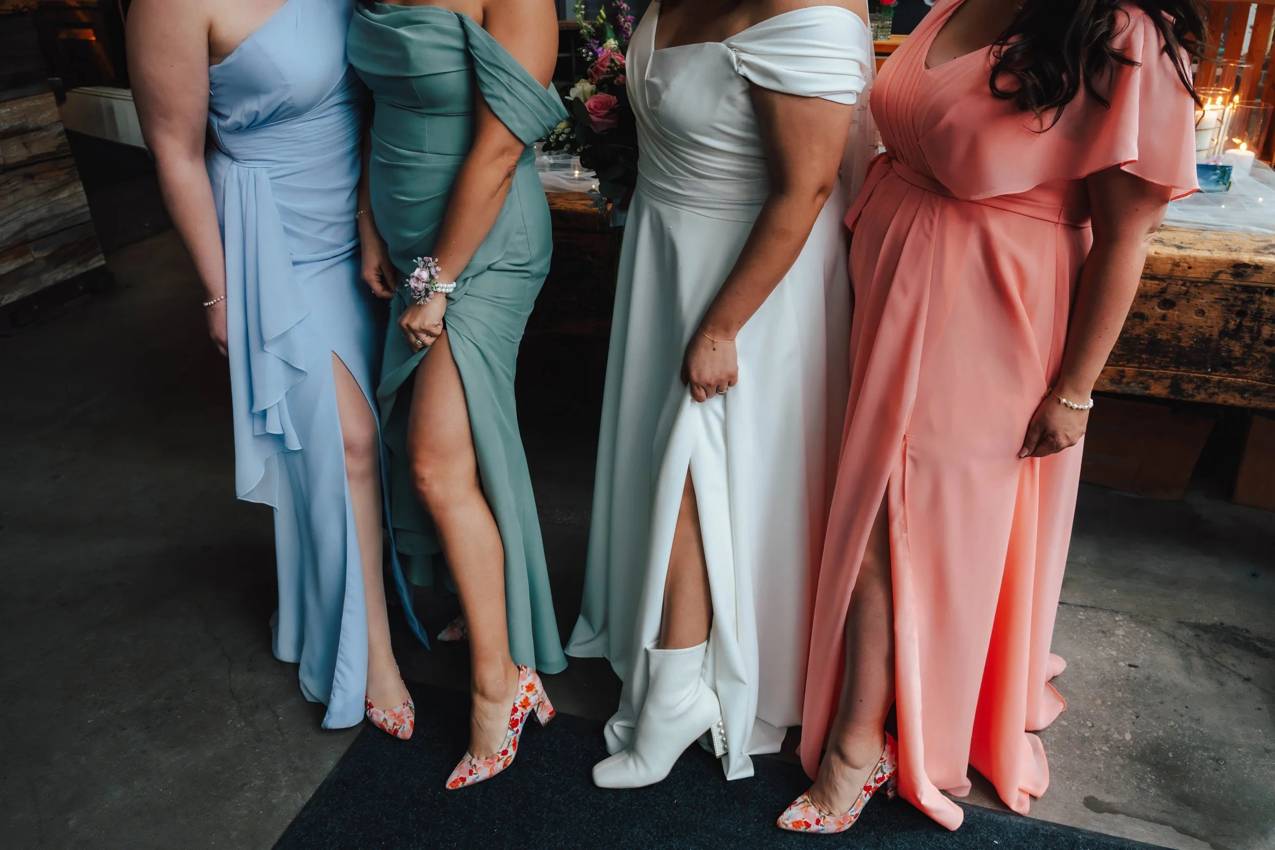 Four women in formal dresses standing indoors, showing their legs through high slits in their dresses, with floral or patterned shoes.