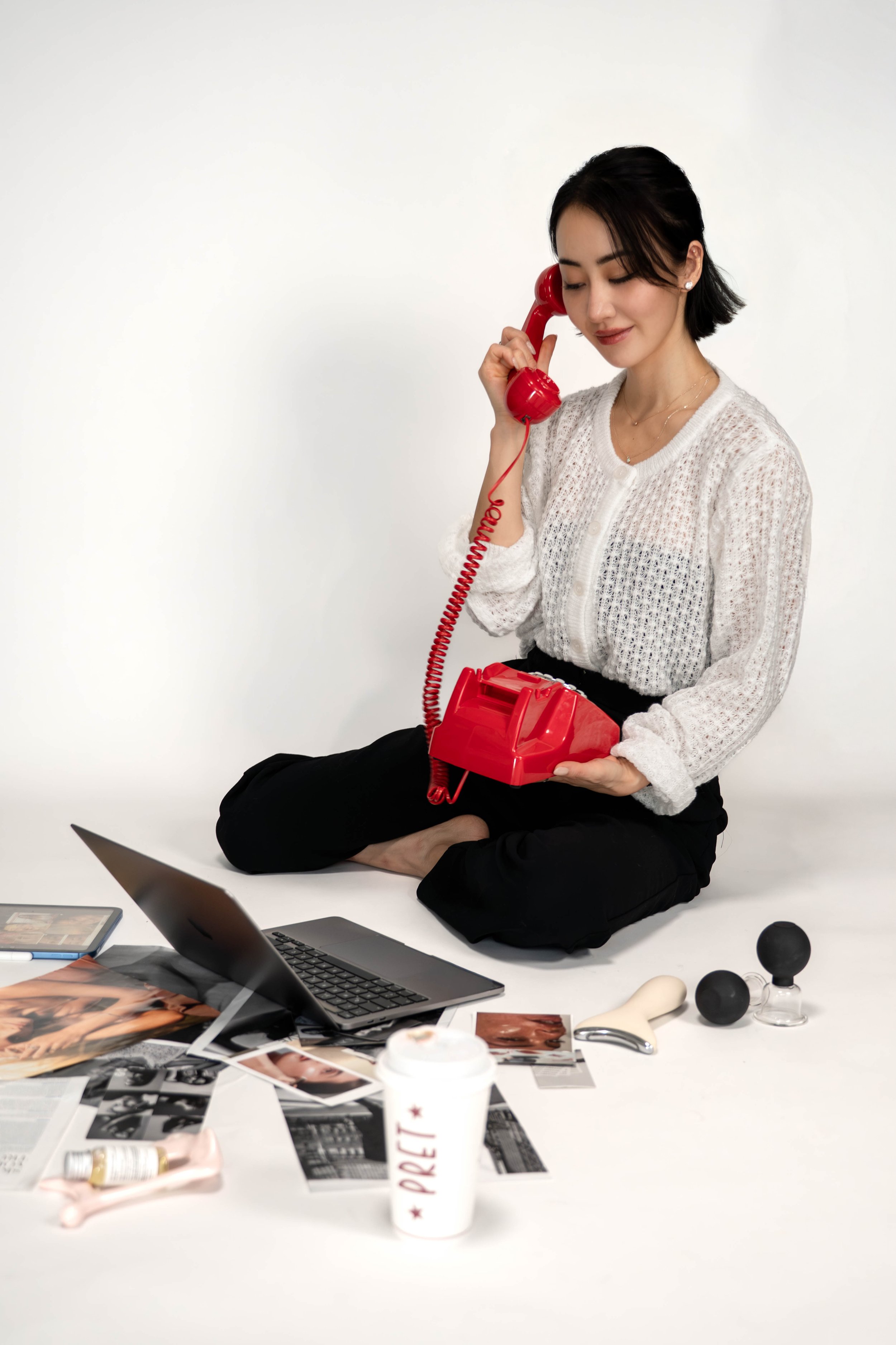 Woman sitting cross-legged on the floor, talking on a red rotary phone, surrounded by magazines, a laptop, and beauty products.