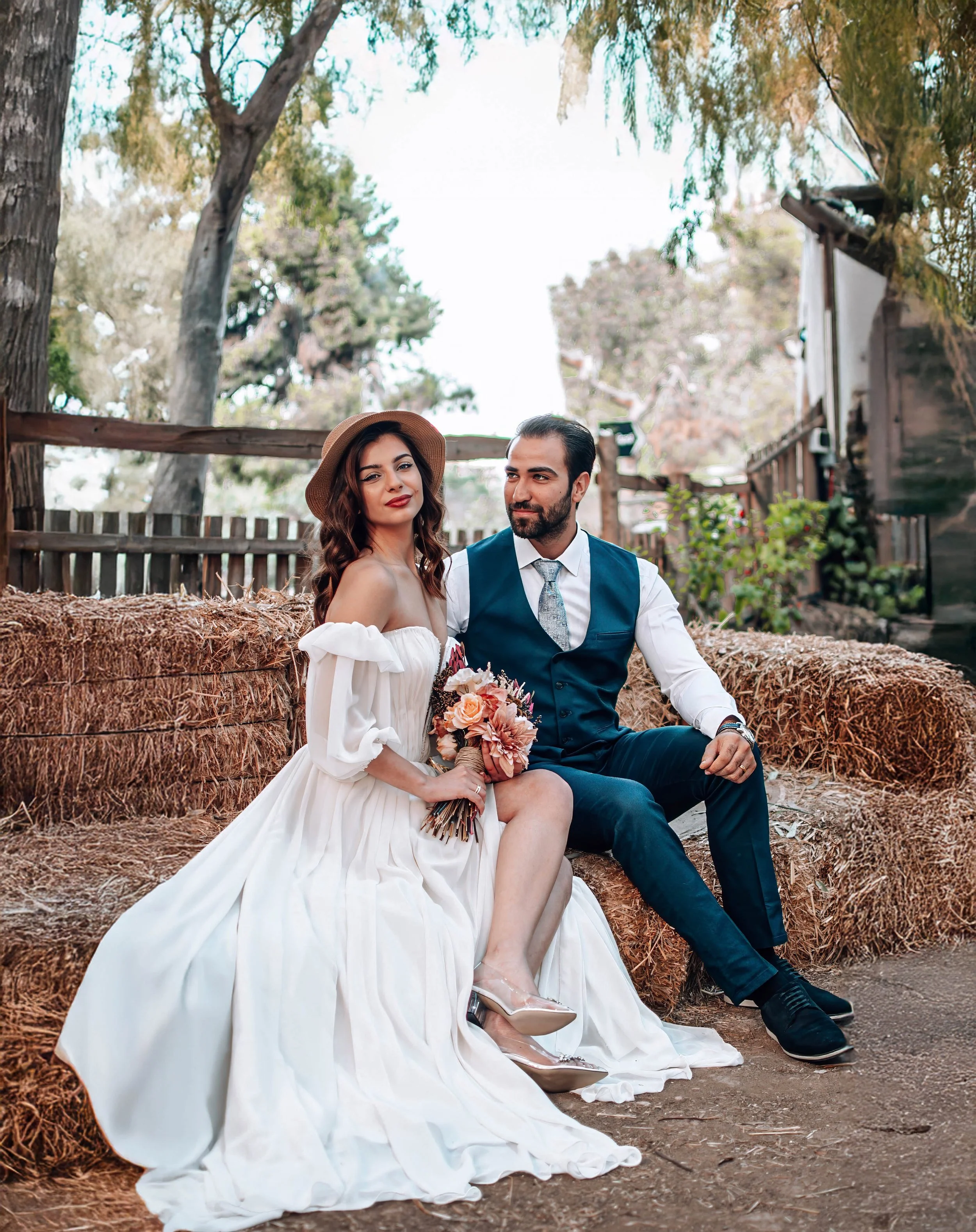 A woman in a white off-shoulder dress and a man in a navy suit sitting on hay bales outdoors, holding a bouquet of flowers, surrounded by trees and rustic wooden fencing.