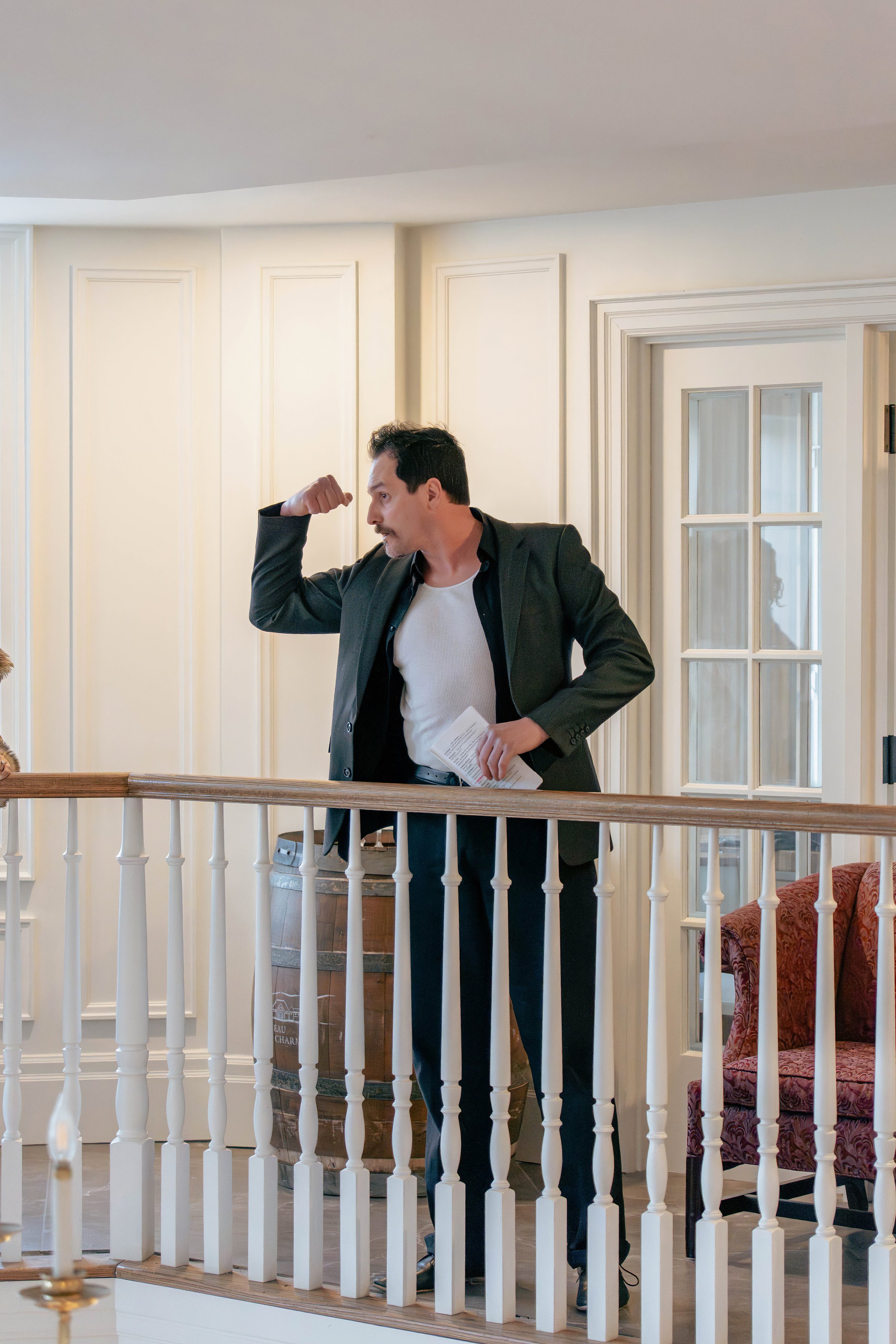 Man in a black blazer flexing his arm muscle on staircase railing inside a house.