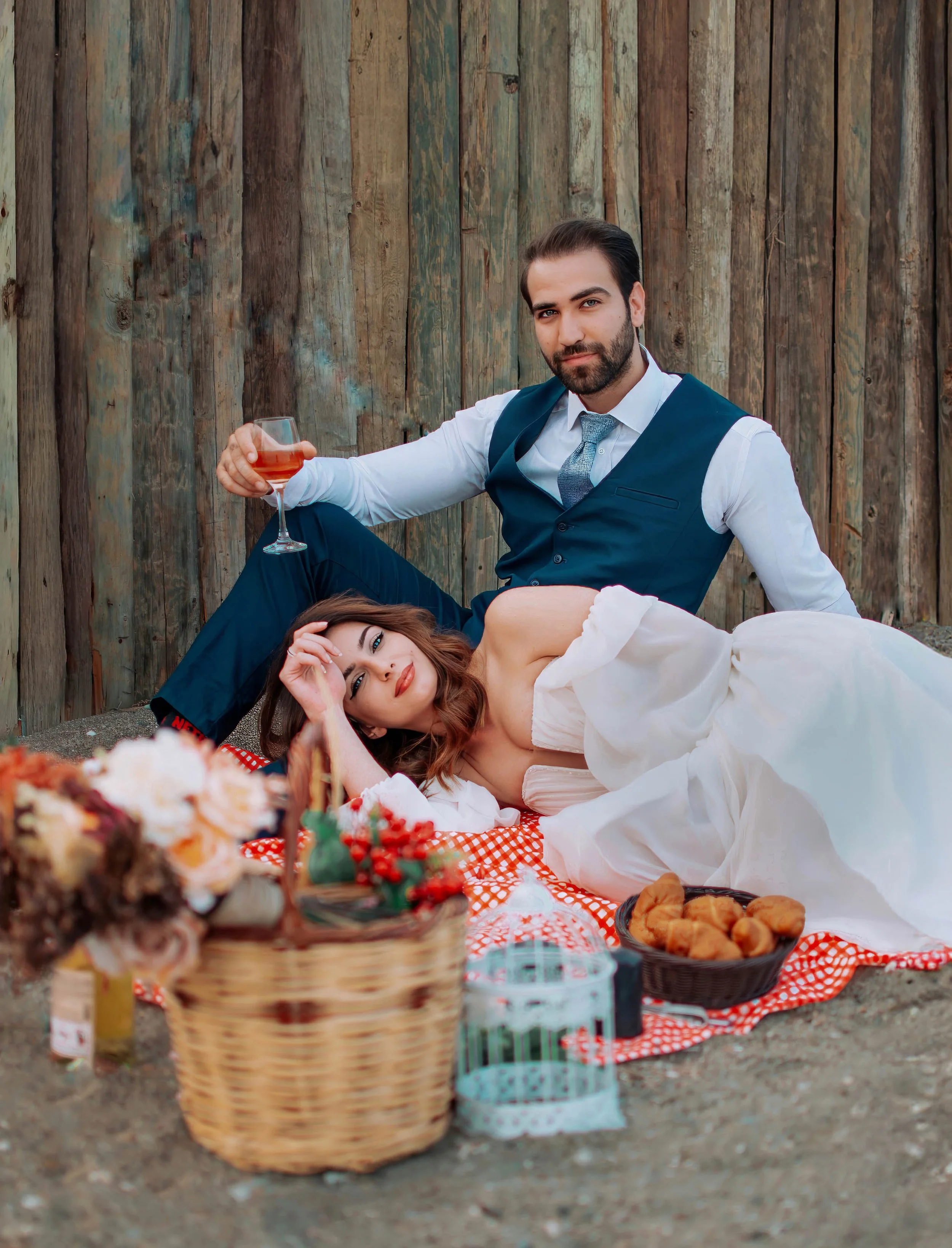 A man and woman having a picnic outdoors against a wooden fence. The man is sitting with a wine glass, dressed in a suit. The woman is lying on a blanket in a white dress, resting her head on her hand. There is a picnic basket, a bowl of croissants, 