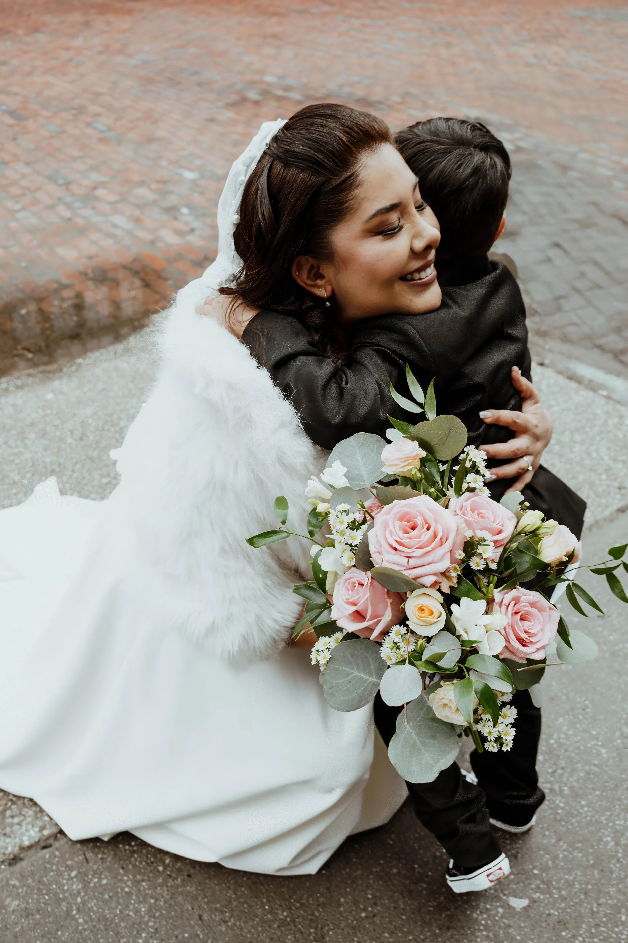 A bride with a white gown and white fur shawl hugging a young boy dressed in black outside on a brick and concrete sidewalk. The bride holds a large bouquet of pink roses, white flowers, and green leaves.