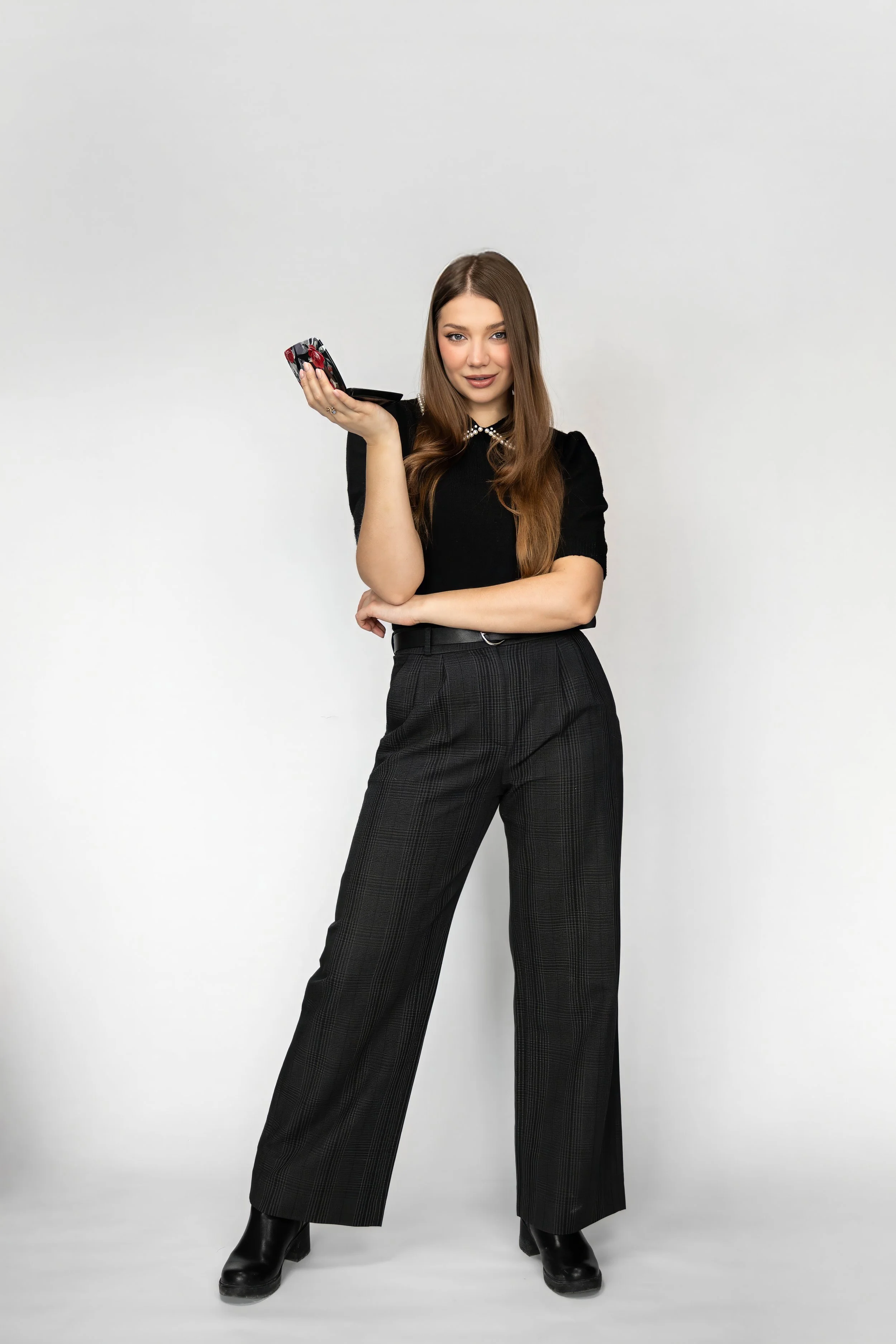 A young woman with long brown hair, wearing a black top, plaid dark pants, and black boots, is standing against a plain white background, holding a makeup palette in her right hand and looking at the camera.