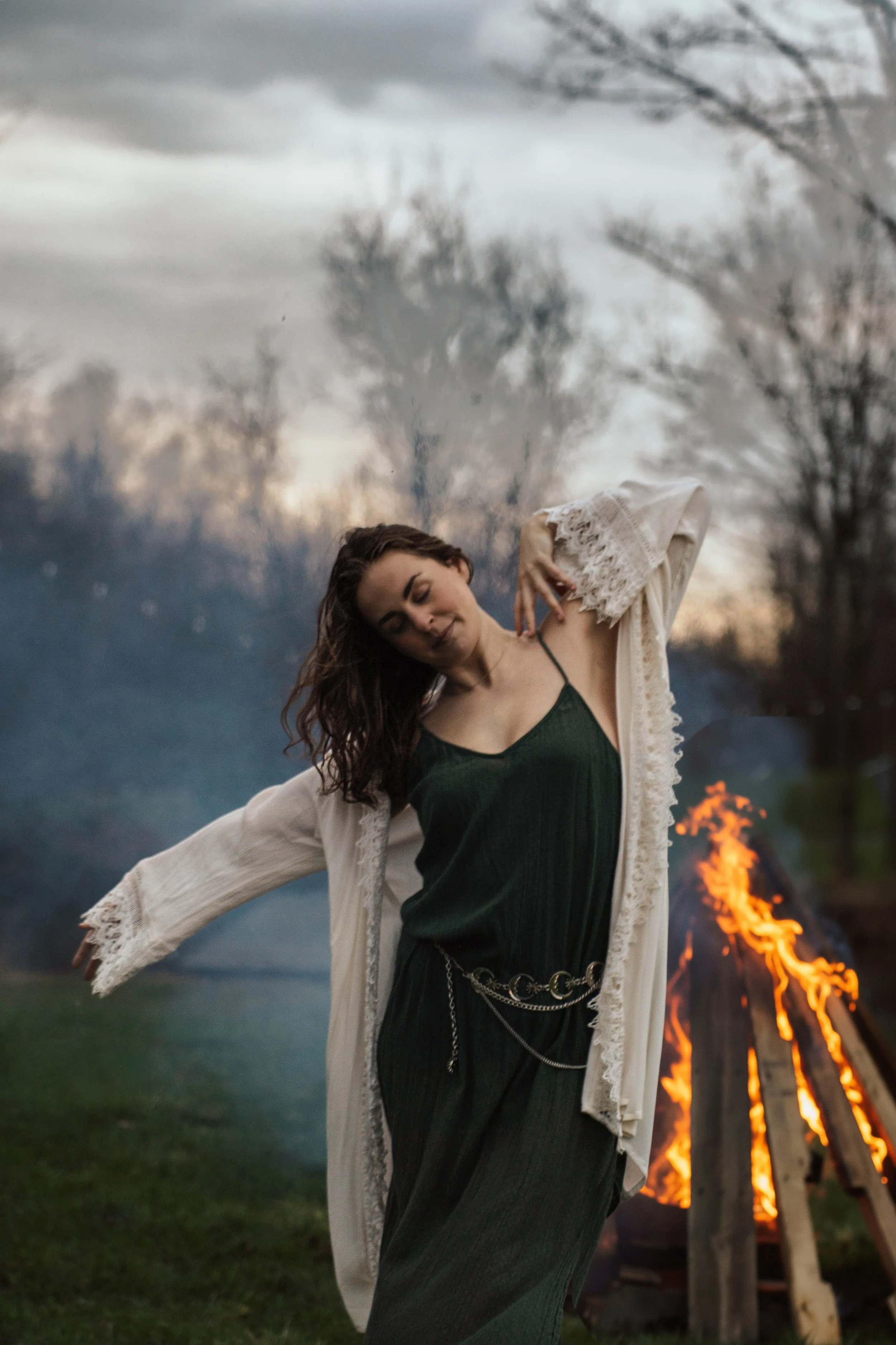 A woman in a green dress and white lace robe dancing near a bonfire outdoors during dusk.