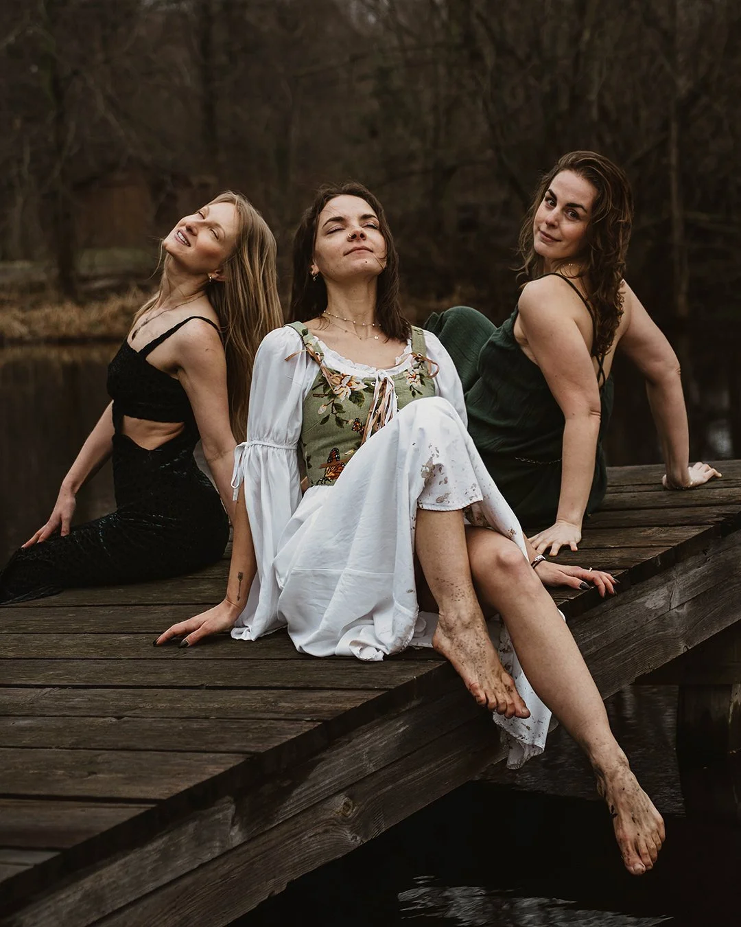 Three women sitting on a wooden dock near a body of water in a natural outdoor setting, with neutral expressions and casual poses.