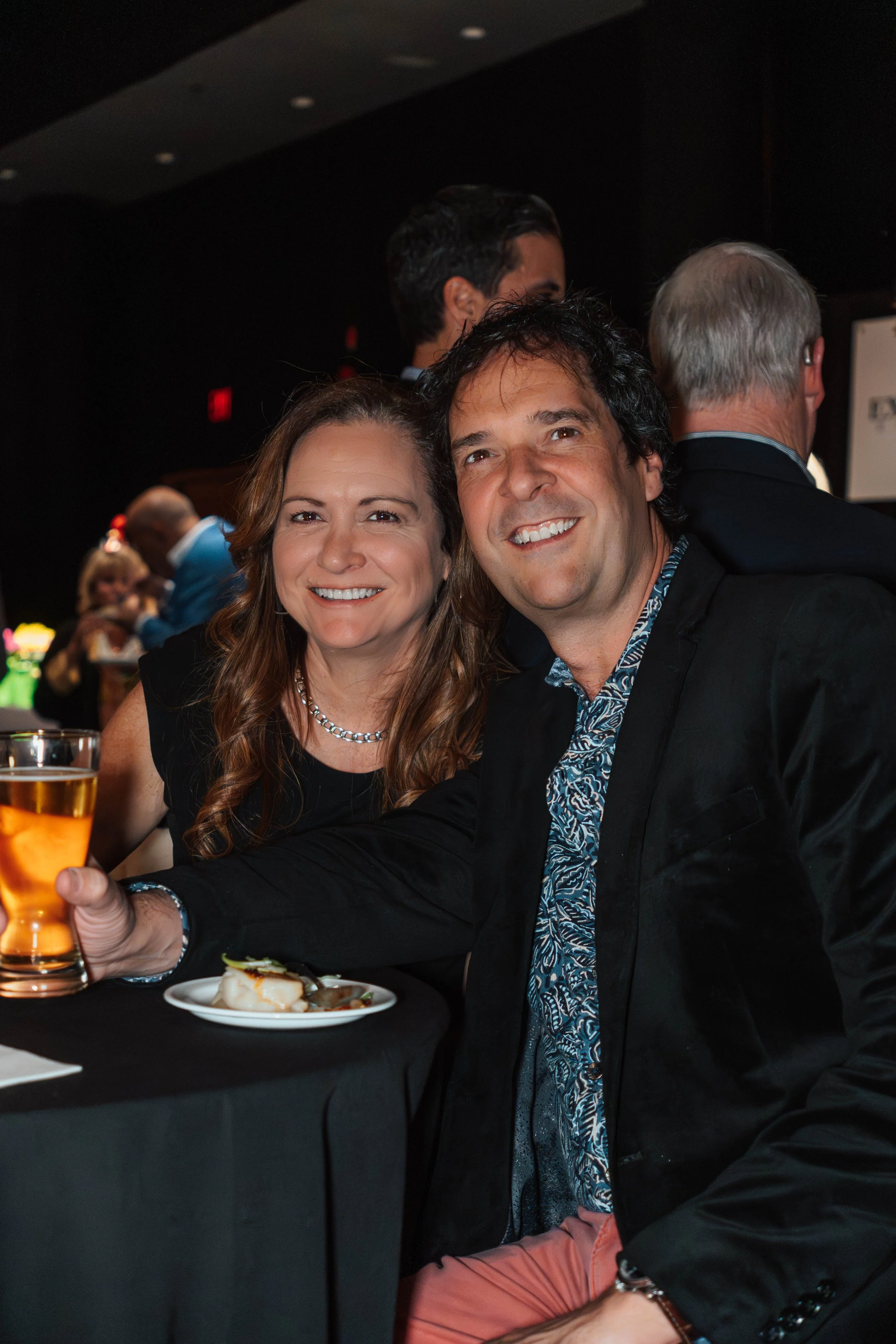 A smiling woman with wavy brown hair and a pearl necklace sits next to a smiling man with dark hair and a patterned blue shirt, both holding drinks, at a social event.