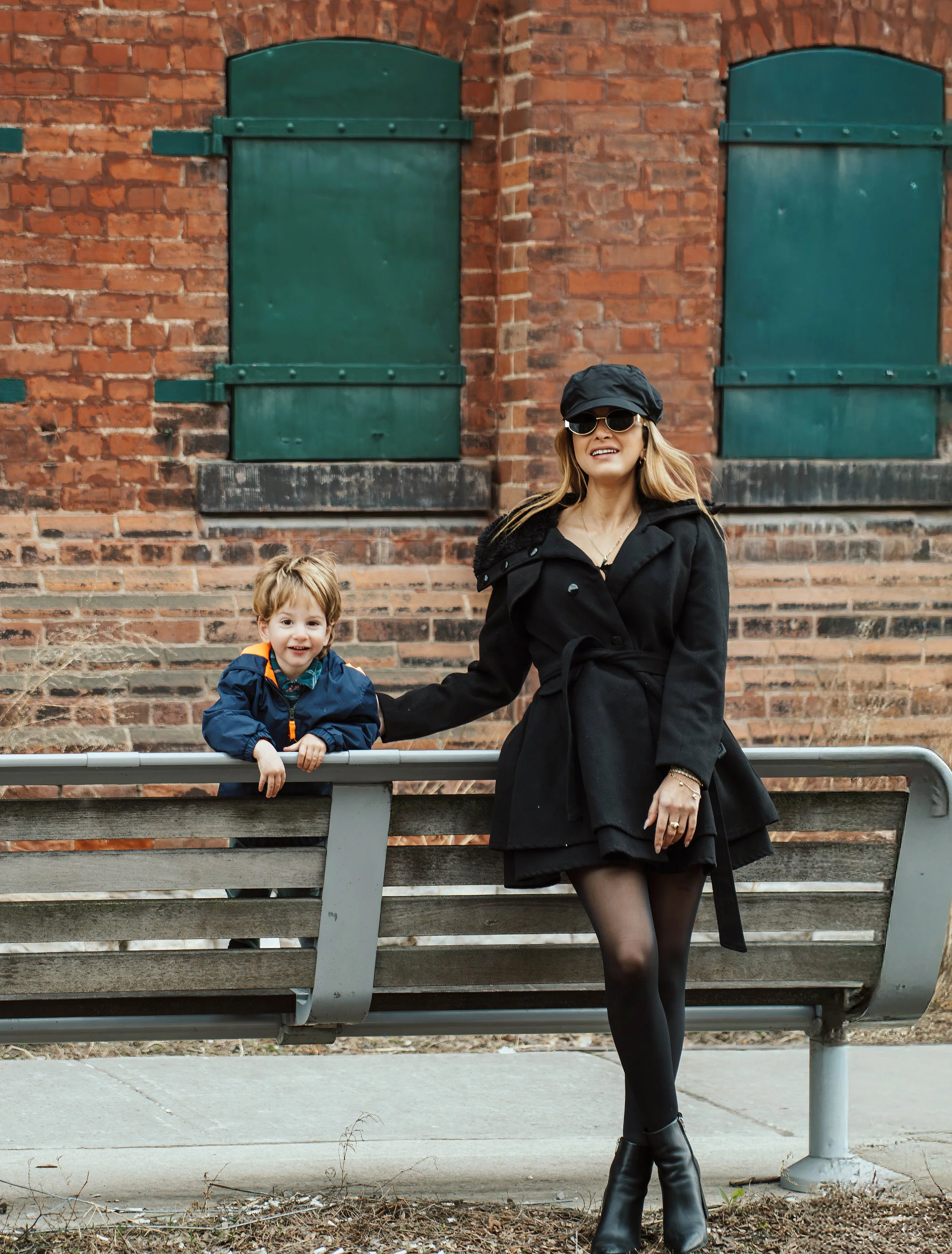 A woman dressed in black with boots, sunglasses, and a black hat, standing next to a small child in a blue and orange jacket, both leaning on a park bench in front of a brick wall with green shutters.