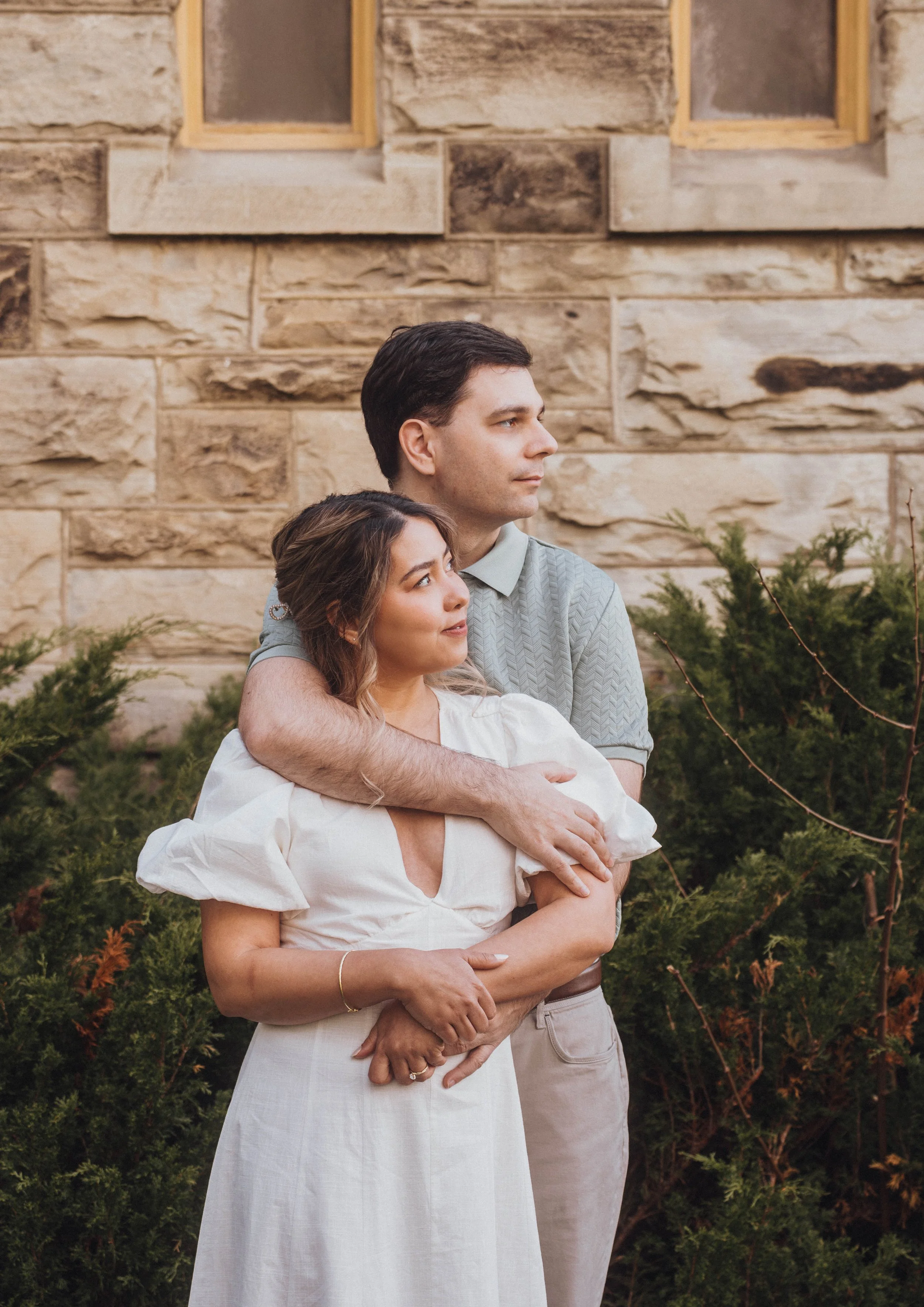A young man and woman standing close together outdoors with a stone wall background, the man hugging the woman from behind, both looking off into the distance.