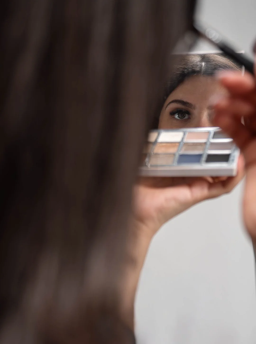 A woman applying eyeshadow using a makeup palette, reflected in a mirror.