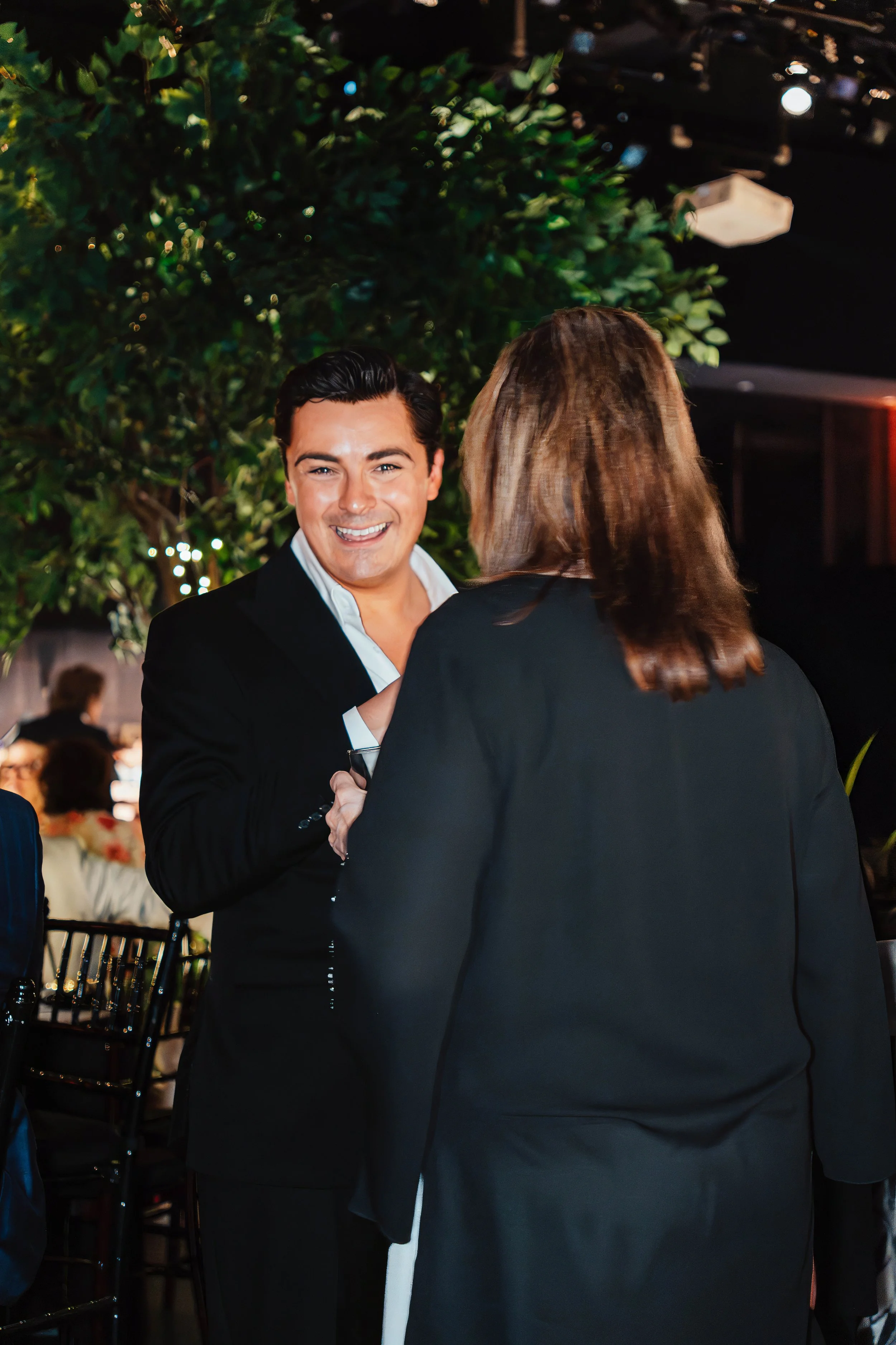 A man smiling and talking to a woman at a social event, with greenery and other people seated at tables in the background.