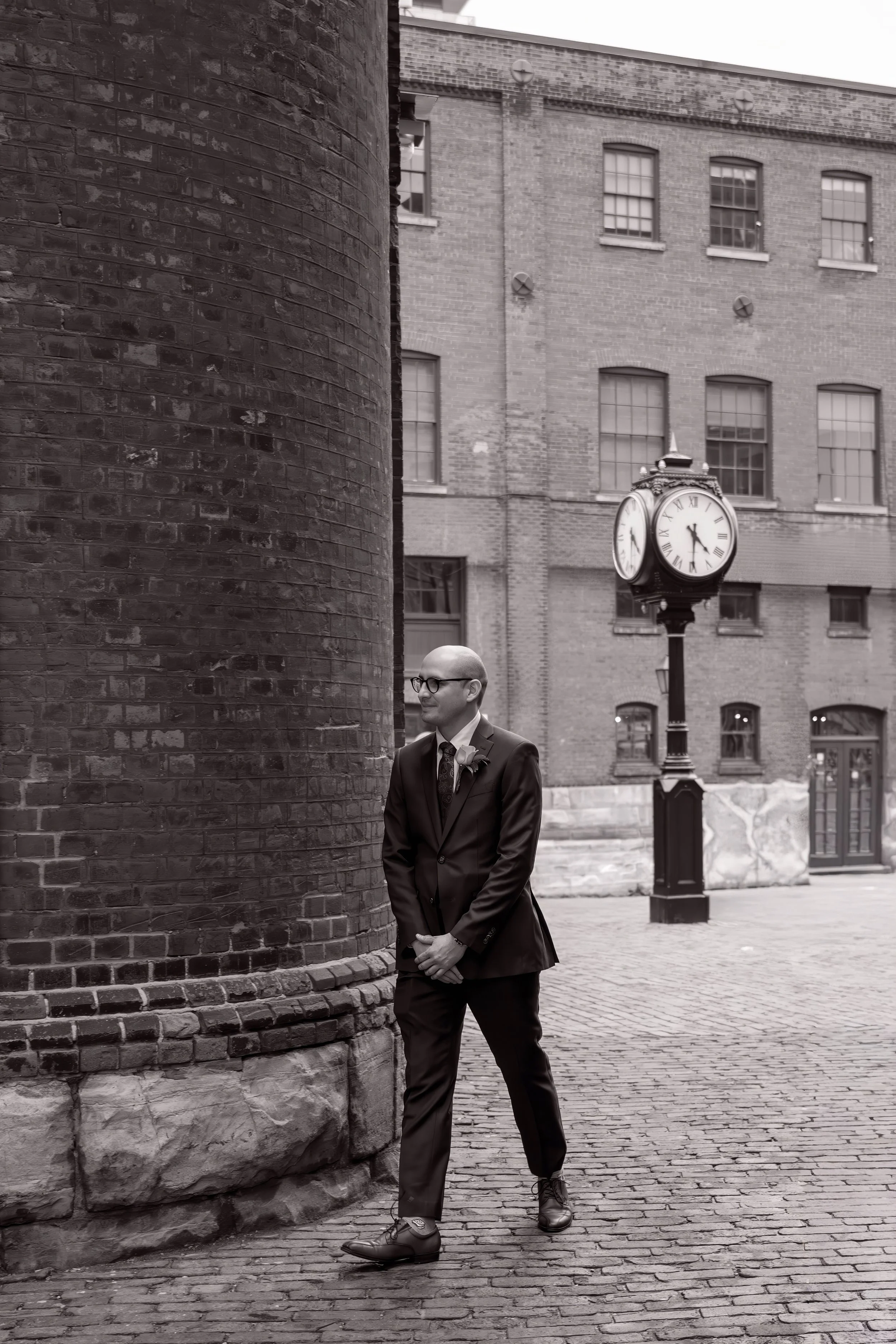 Black and white groom portrait in Toronto’s Distillery District with historic brick architecture, vintage clock, and cinematic documentary wedding photography style.