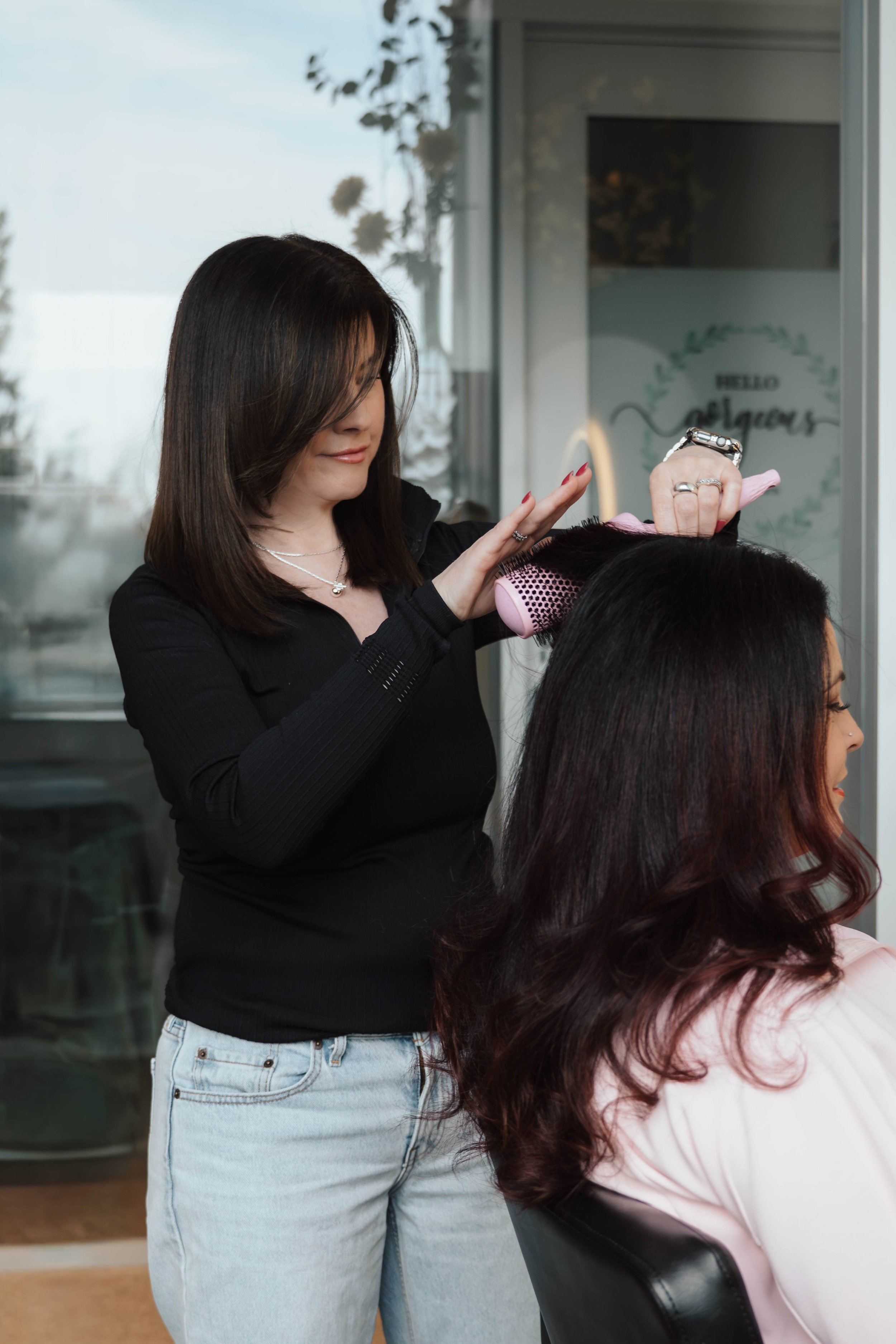 A hairstylist drying a woman's long dark hair with a pink hairbrush near a window in a salon.