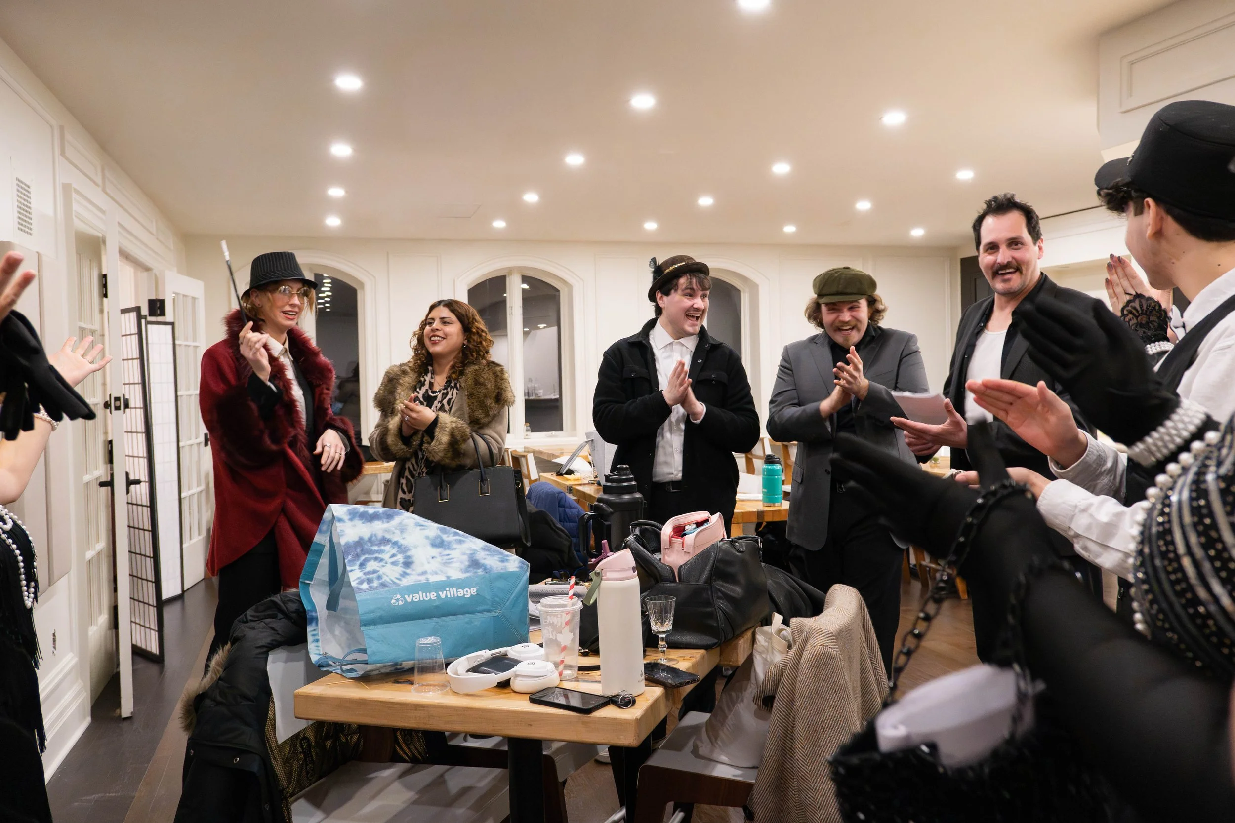 Group of people dressed in costumes, smiling and clapping in a festive indoor setting with a table full of personal items and shopping bags.