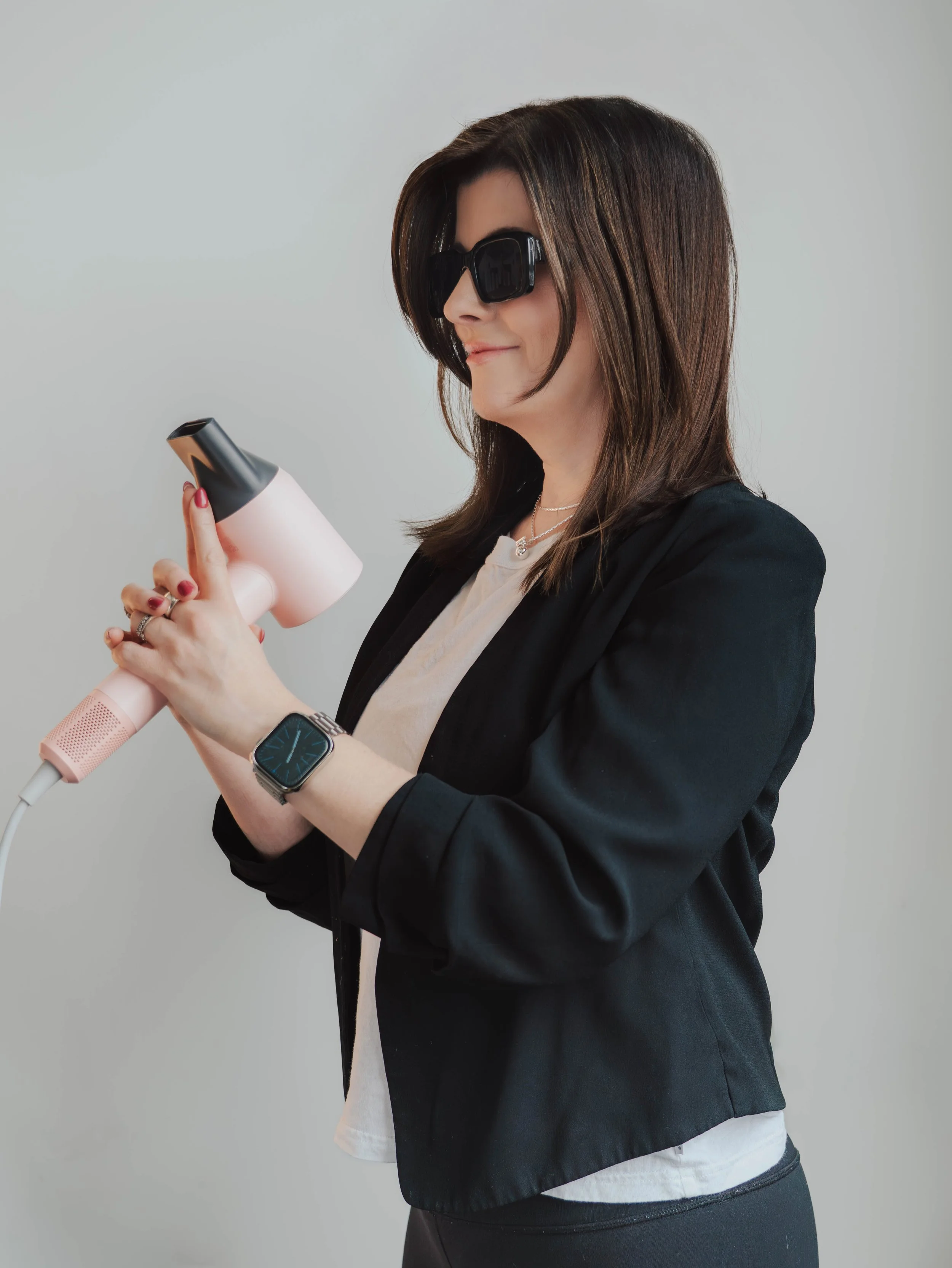 Woman with long brown hair wearing black sunglasses, black blazer, and a smartwatch, holding a pink hair dryer and styling tool.