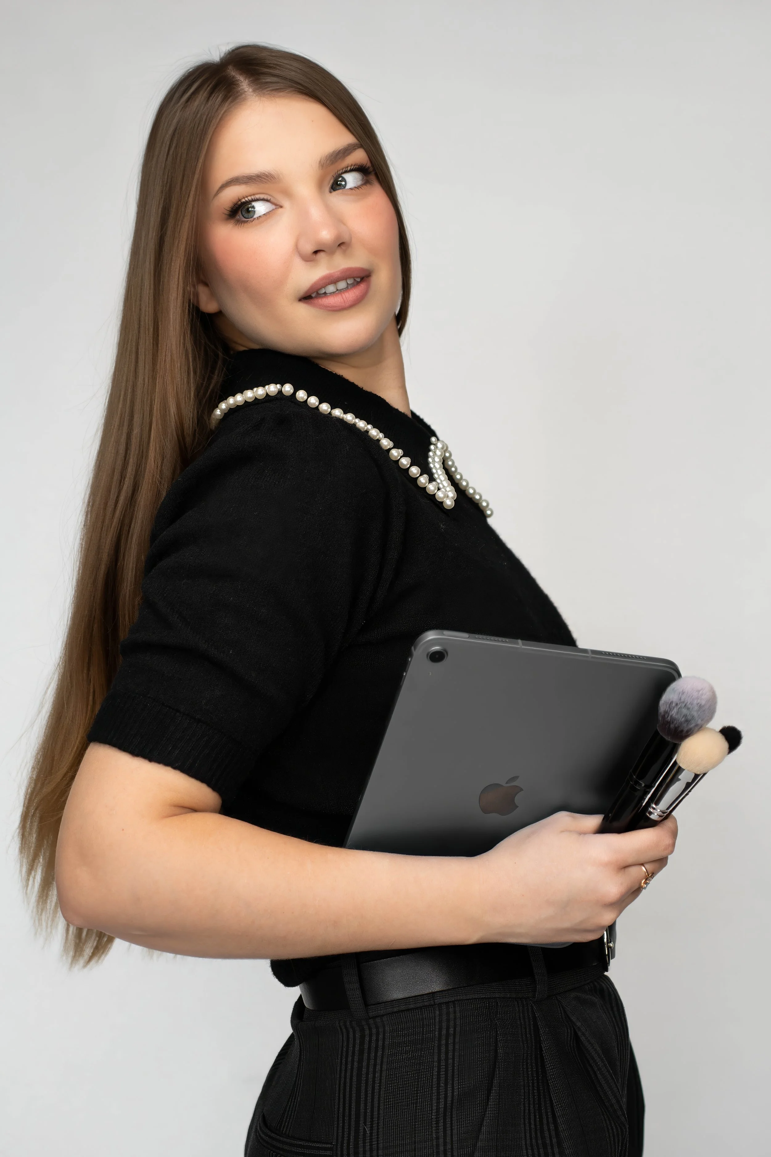 A woman with long brown hair and makeup holding a gray iPad and makeup brushes, wearing a black top with pearl embellishments on the collar in front of a plain gray background.