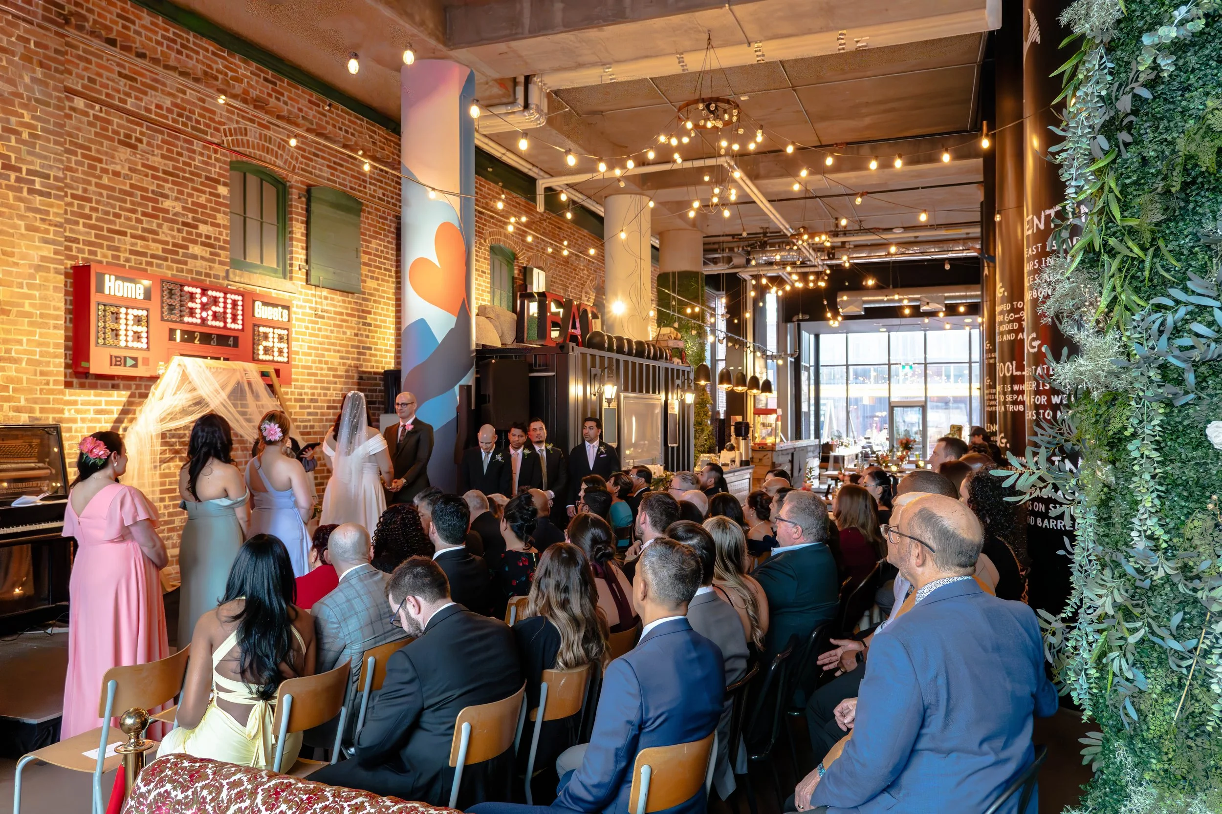 A wedding ceremony taking place inside a stylish venue with brick walls, string lights, and large windows; guests are seated watching the couple exchange vows, with bridesmaids and groomsmen standing nearby.