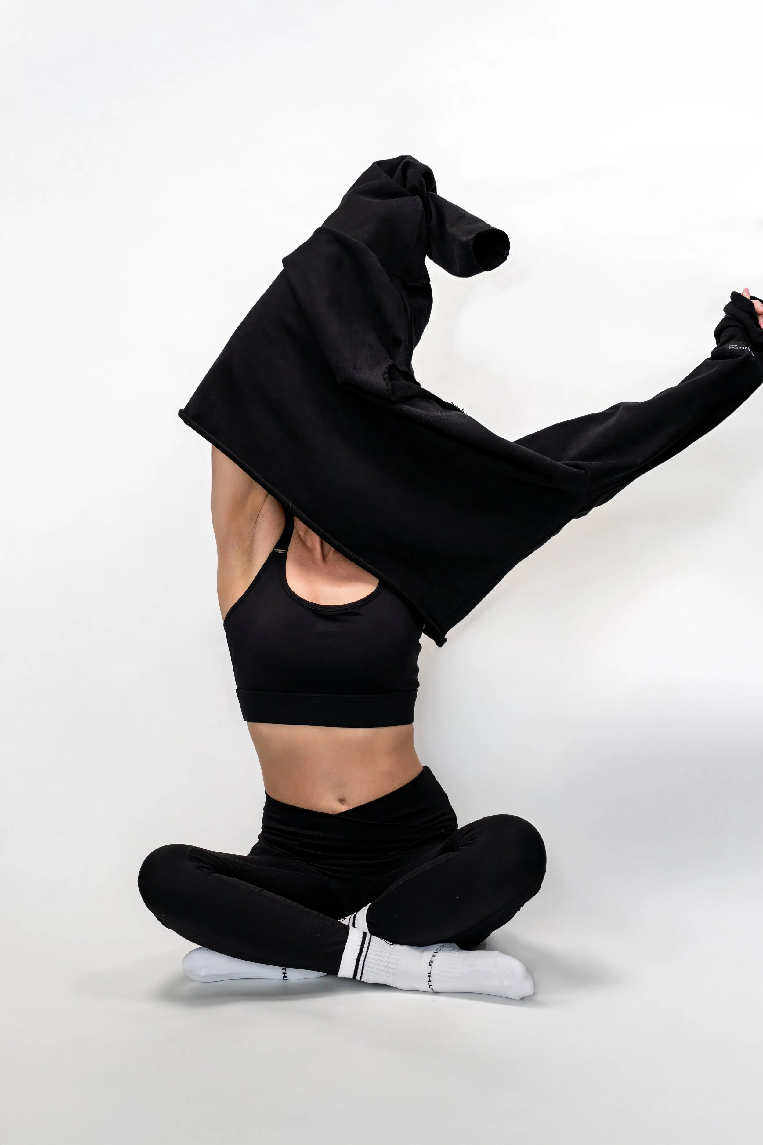 Person in black athletic wear sitting cross-legged on the floor, raising a black hoodie over their head with arms extended.