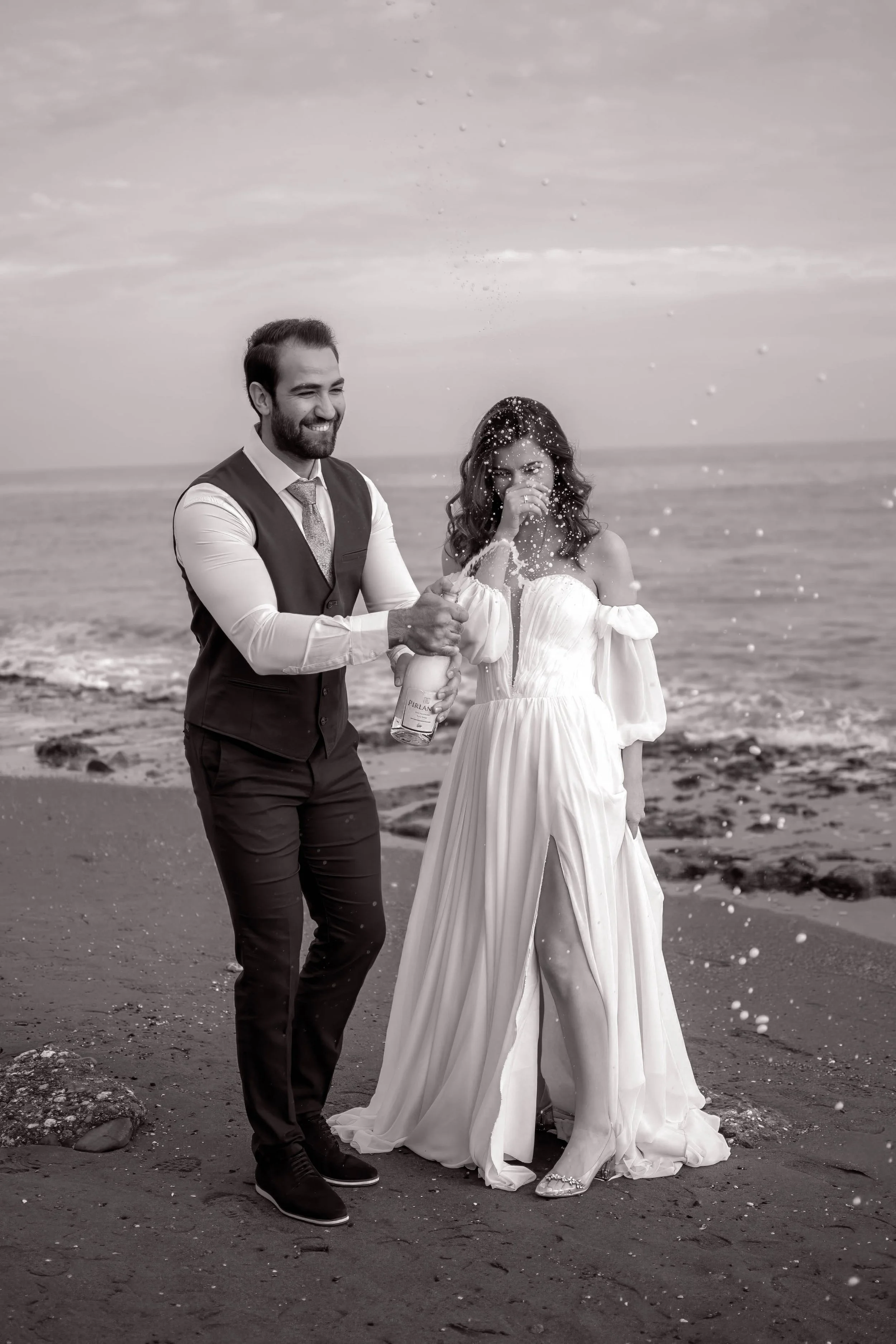 A couple in wedding attire celebrating on the beach, with the man opening a champagne bottle and champagne spraying out, as the woman holds her nose and smiles.