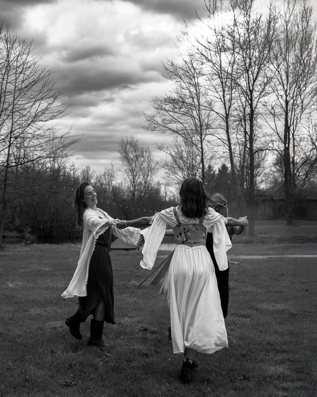 Three women dancing outdoors on a grassy area with leafless trees and clouds in the sky, black and white photo.
