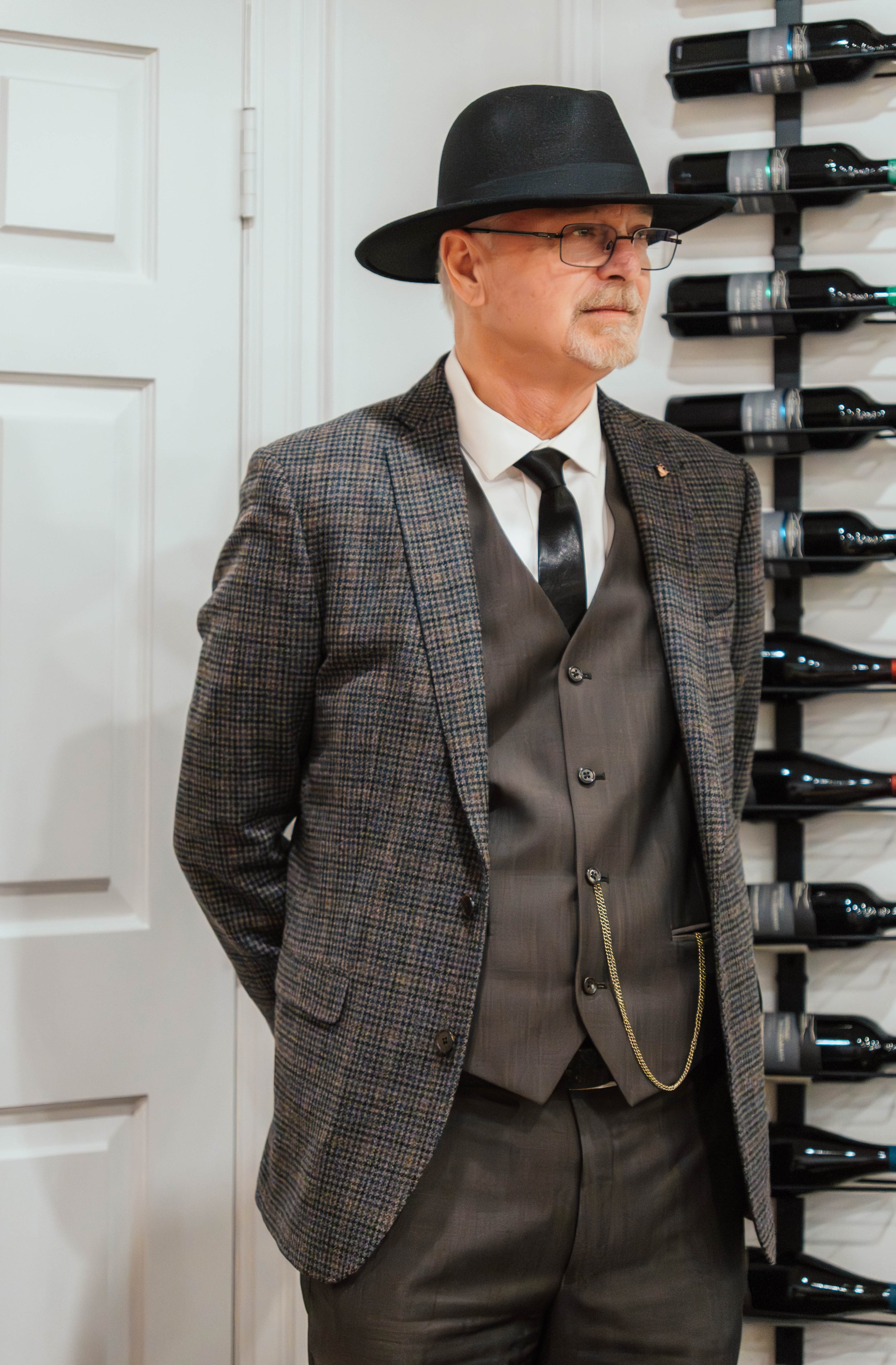 An older man dressed in a vintage-style suit and hat, standing next to a wine rack.