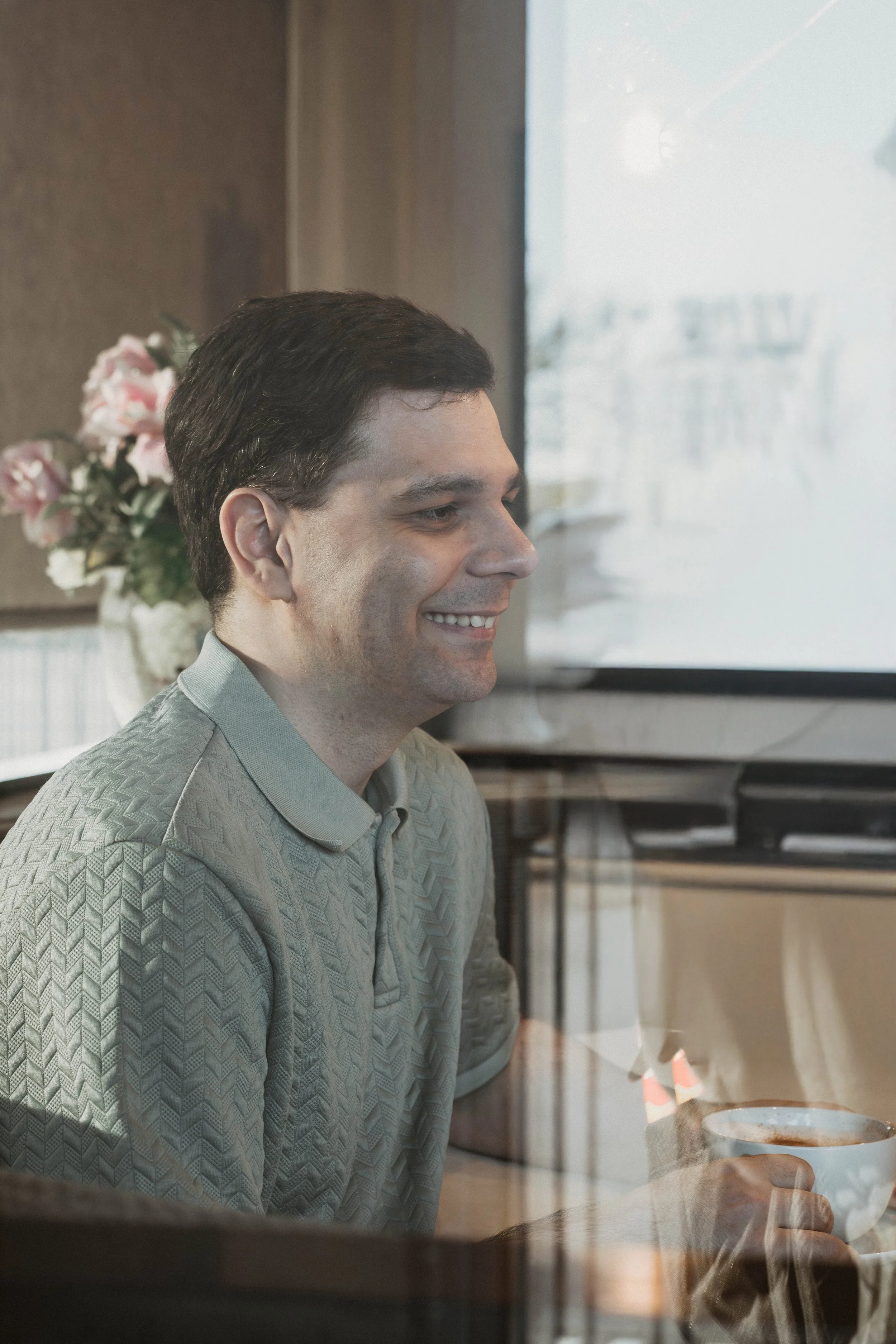 A man with dark hair sitting at a table, smiling, with a drink in his hand. In the background, a window and a bouquet of pink and white flowers are visible.