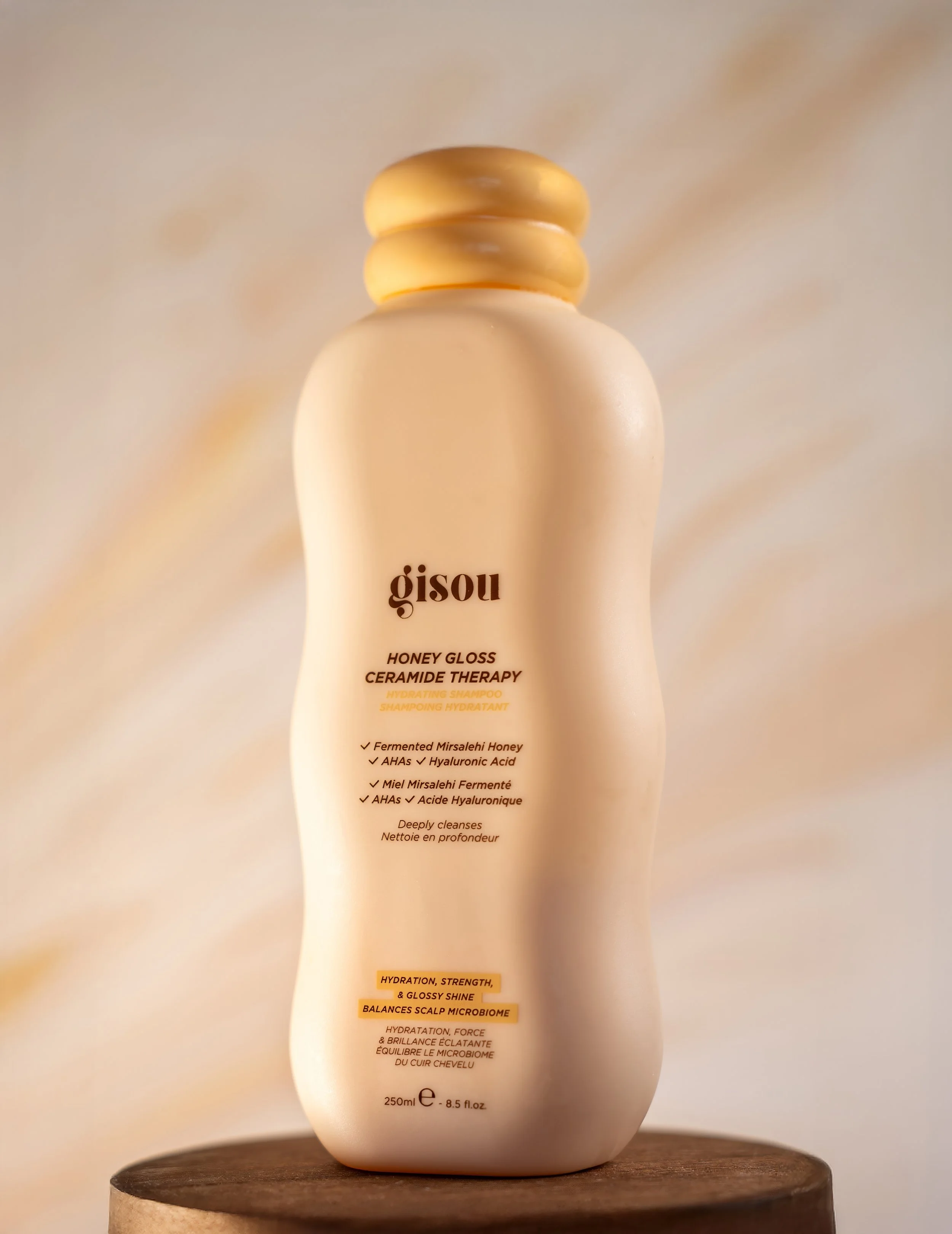 A beige bottle of Gisou Honey Gloss Ceramide Therapy Hydrating Shampoo with a rounded top, placed on a wooden surface with a soft, neutral-colored background.