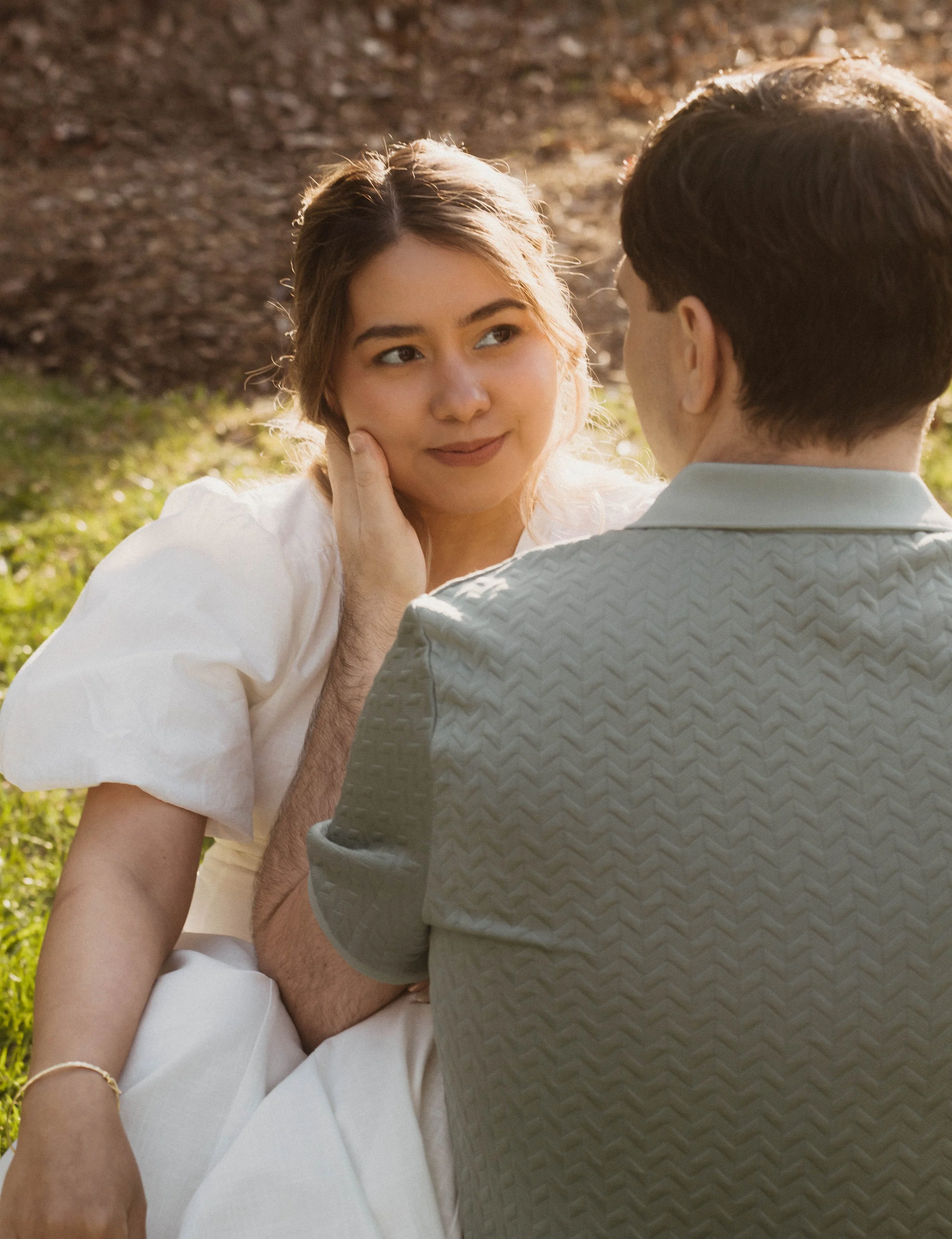 A young woman with light brown hair and fair skin looking at a young man with dark brown hair and fair skin, outdoors in a park, with soft sunlight and trees in the background.